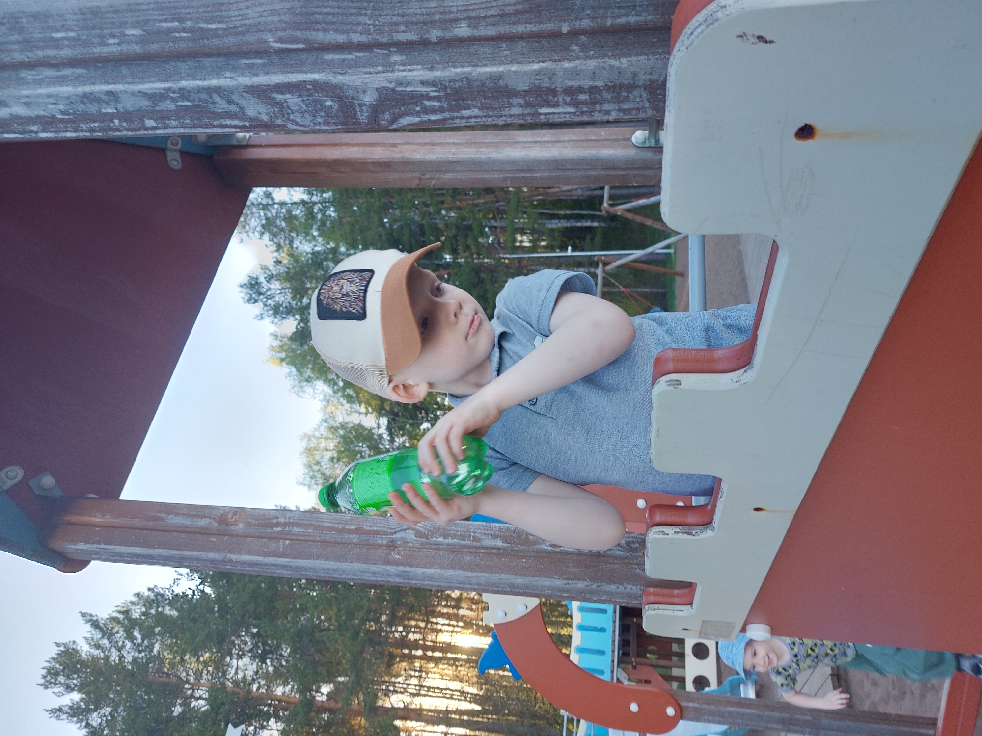 A young boy, wearing a tan and white baseball cap with a lion insignia, and a light blue collared shirt, is holding a green plastic bottle on a playground. The playground structure appears to be made of painted wood and metal, with sections of red, white, blue, and tan. Behind the boy, there are trees visible against the sky, indicating an outdoor location, probably at dusk or dawn. Another child is visible in the bottom right corner wearing a patterned shirt and a blue hat. Based on prior knowledge, this image was taken in the city of Chaikovsky, Russia.