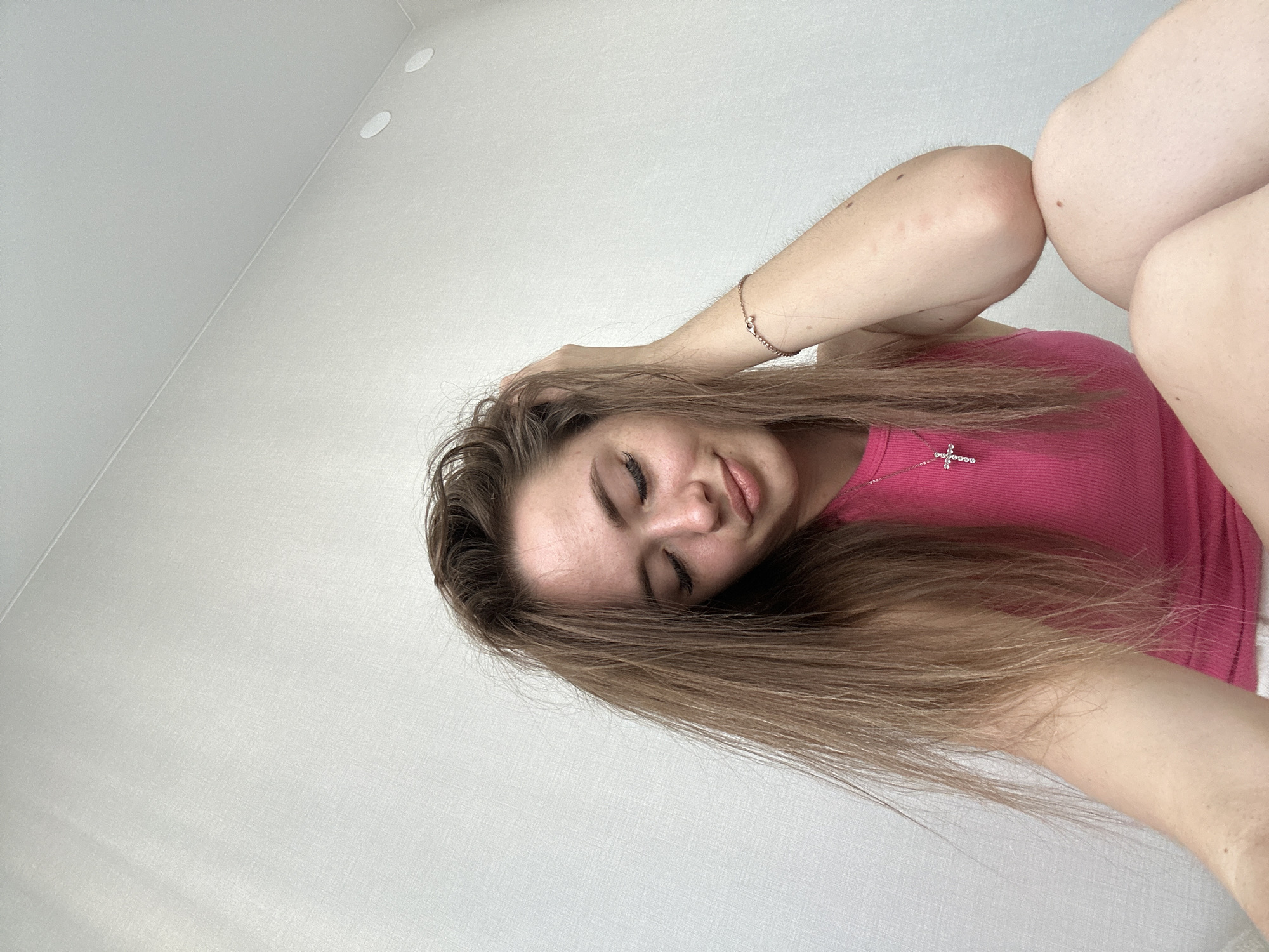 A young woman is the central subject of this low-angle, upward-facing photograph. She appears to be lying down or leaning back, looking towards the camera with her eyes closed and a soft, relaxed expression on her face. Her lips are slightly parted. She has long, straight light brown hair that cascades around her head. She is wearing a bright fuchsia or pink ribbed tank top. A delicate silver or white gold chain necklace with a small, sparkly cross pendant adorns her neck, and a thin, light-colored bracelet is visible on her left wrist. One of her arms is bent, with her hand gently touching her hair. Parts of her bare arms and legs are also visible in the frame, suggesting a relaxed pose. The background consists of plain, light-colored, possibly off-white or very light grey walls and a ceiling, which appear to have a subtle texture. Two small, circular, light-colored fixtures, possibly recessed lights or vents, are visible on the ceiling in the upper left area of the image. The lighting is soft and even, characteristic of an indoor setting. There is no visible text.