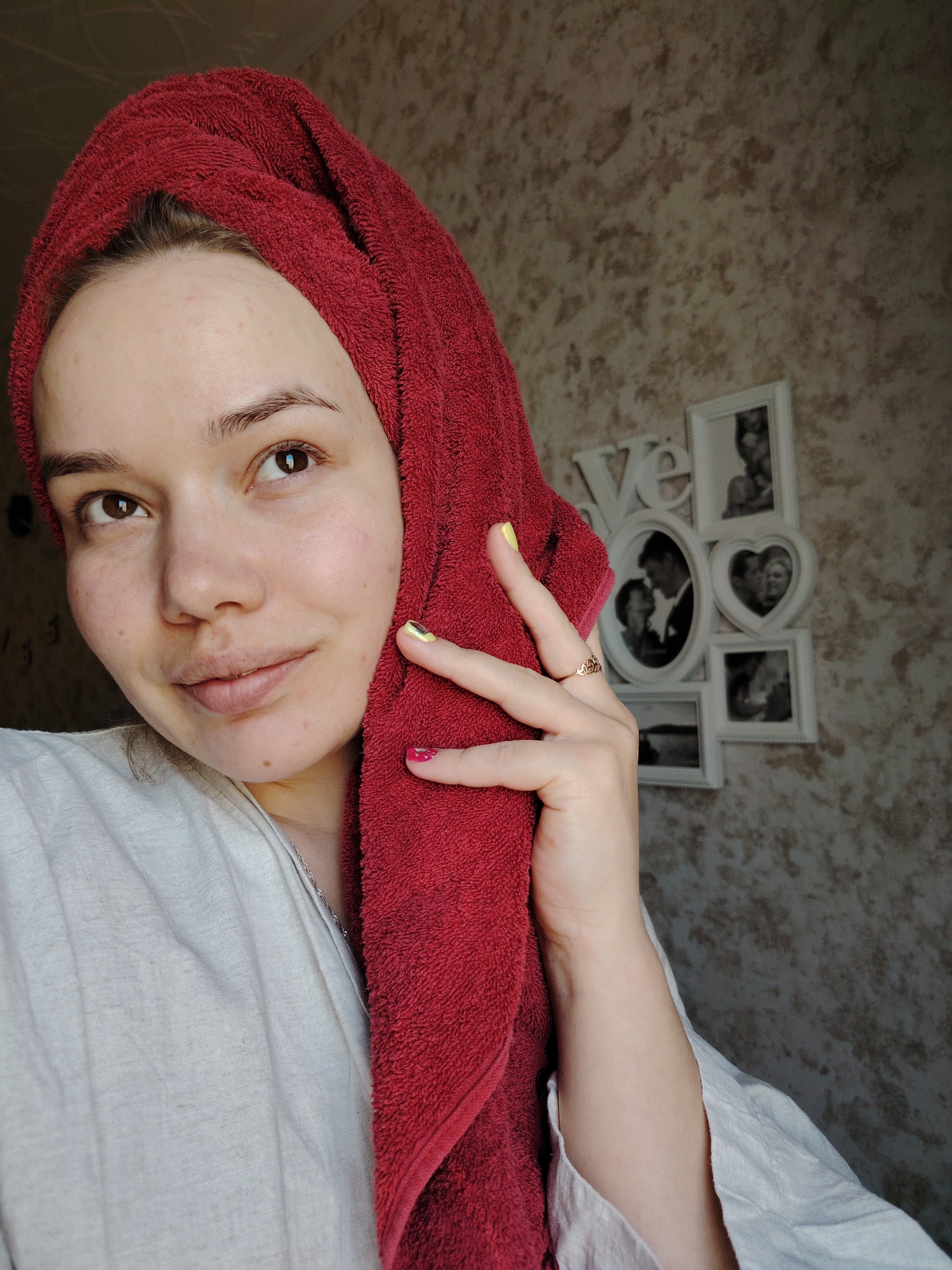 An image taken indoors, presumably in the city of Марковский, Russia, depicts a female individual in a medium shot. The subject, a young adult female with fair skin, brown eyes, and brown eyebrows, is looking directly towards the camera with a faint smile. Her hair is wrapped in a dark red terrycloth towel, and she is wearing a light beige or off-white collared garment. A thin silver-colored chain is visible around her neck. Her left hand is raised, with fingers slightly splayed, resting against the towel near her left cheek. The index finger of her left hand displays yellow nail polish, and the middle finger displays pink nail polish. A thin gold-colored ring, possibly adorned with heart shapes, is visible on her left ring finger. The background consists of a textured wall, likely wallpaper, in mottled shades of brown and beige. To the upper right, a wall decoration features multiple white-framed photographs in various shapes (rectangular, oval, heart), arranged around the letters "L V E," which appear to be part of the word "LOVE." The framed photographs contain black and white images, predominantly depicting couples.