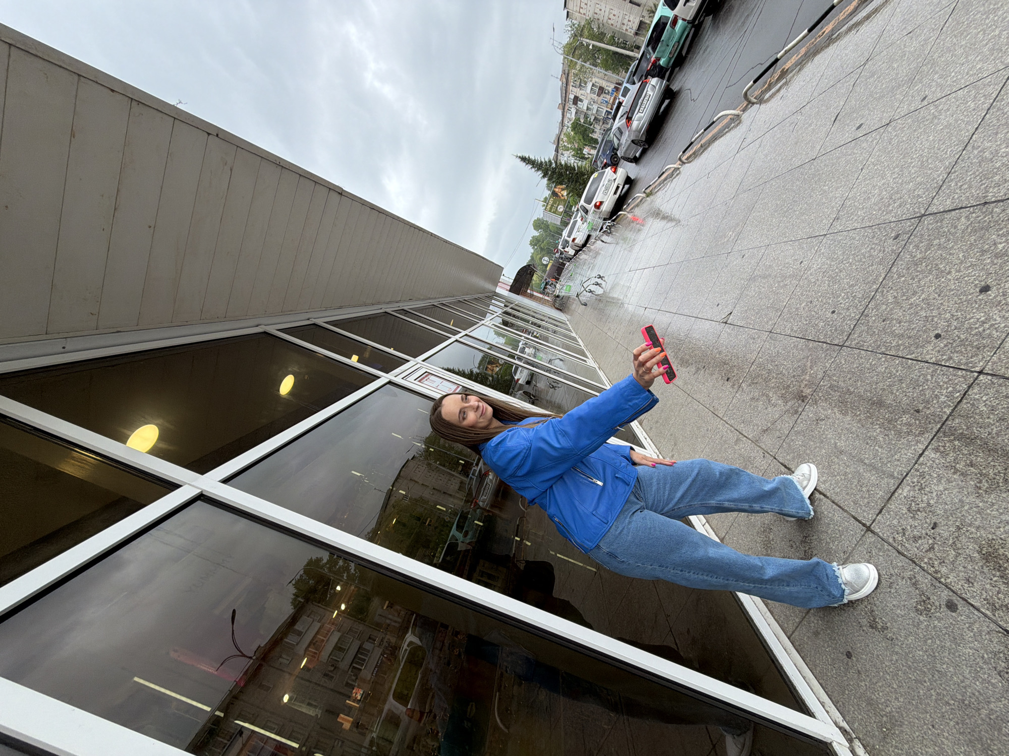 A young woman takes a selfie in front of a commercial building with large windows in Birobidzhan, Russia. She is wearing a blue jacket, blue jeans, and white sneakers. She holds a pink phone in her hand. The building reflects buildings and trees, and the sidewalk is wet. Cars are parked along the curb. The sky is overcast.