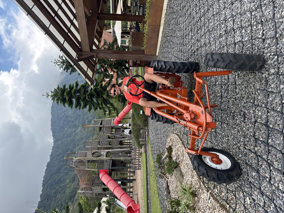 A man in sunglasses and a red t-shirt sits on an orange tractor, holding the steering wheel. The tractor is parked on gravel, with a playground and a mountain in the background. A building with a wooden roof is visible in the upper left corner. The sky is partly cloudy. The orientation of the image is rotated 90 degrees counter-clockwise.
