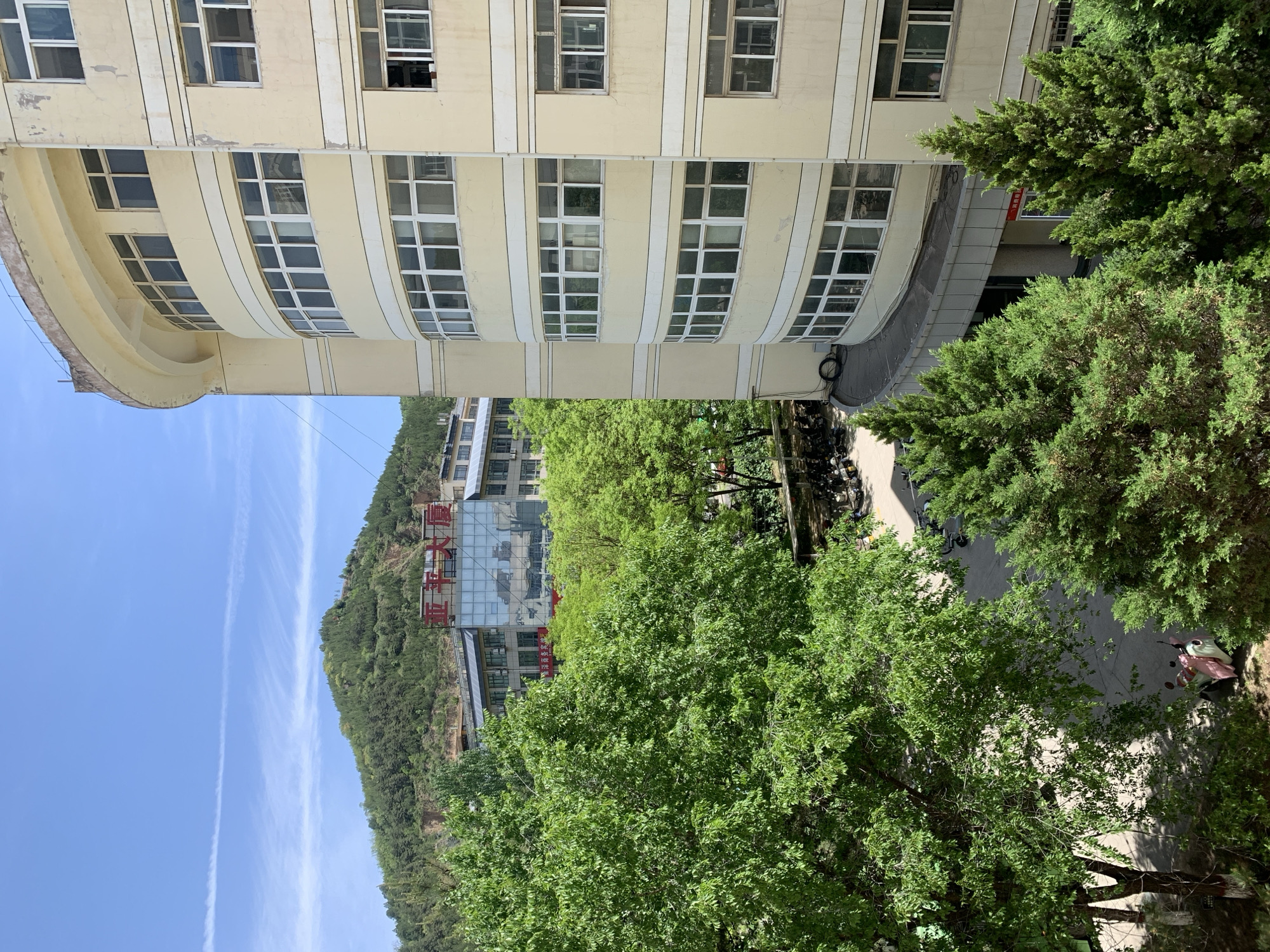 The image depicts a scene in an urban area, likely in Asia given the presence of Chinese characters on a building. The focal point includes a large, multi-story building with a curved facade, light-colored walls, and numerous windows. Adjacent to this building are mature green trees that partially obscure the view of the building in the background, which is nestled against a green hillside. A narrow road or pathway is visible, with parked scooters. The sky is blue with contrails.