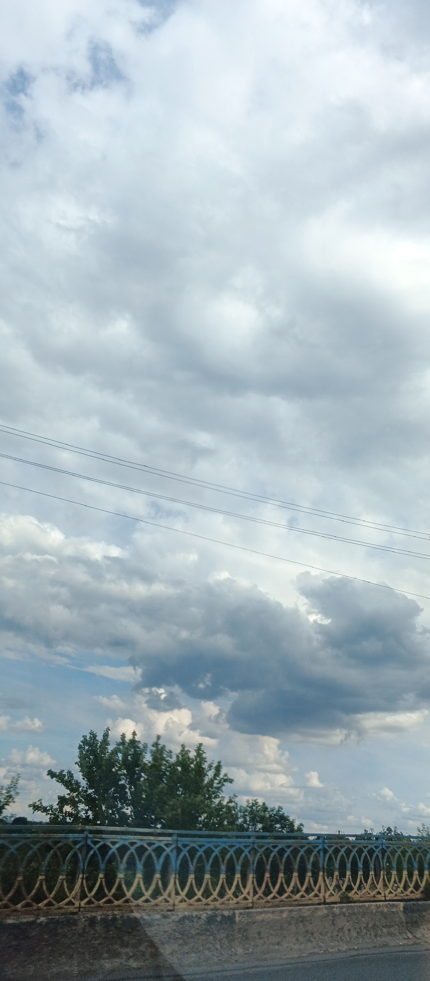 This is a vertical, outdoor, daytime photograph taken from a low angle, likely from a vehicle. The sky dominates the upper two-thirds of the image, filled with a mix of bright white cumulus clouds and darker, grayer clouds, suggesting an overcast or partly cloudy day. Several thin, dark power lines stretch horizontally across the mid-upper portion of the frame.
The lower third of the image features a decorative railing on a bridge or embankment. The railing is ornate, with a repeating pattern of interconnected oval shapes. The colors of the railing appear to be primarily blue and yellow, possibly reminiscent of the Ukrainian flag. Behind the railing, dense green trees are visible, their foliage appearing full and healthy. The very bottom of the image shows a concrete surface, likely part of the bridge or road. The overall impression is a scenic view of nature and infrastructure under a dramatic sky. The location is the city of Smila, Ukraine.