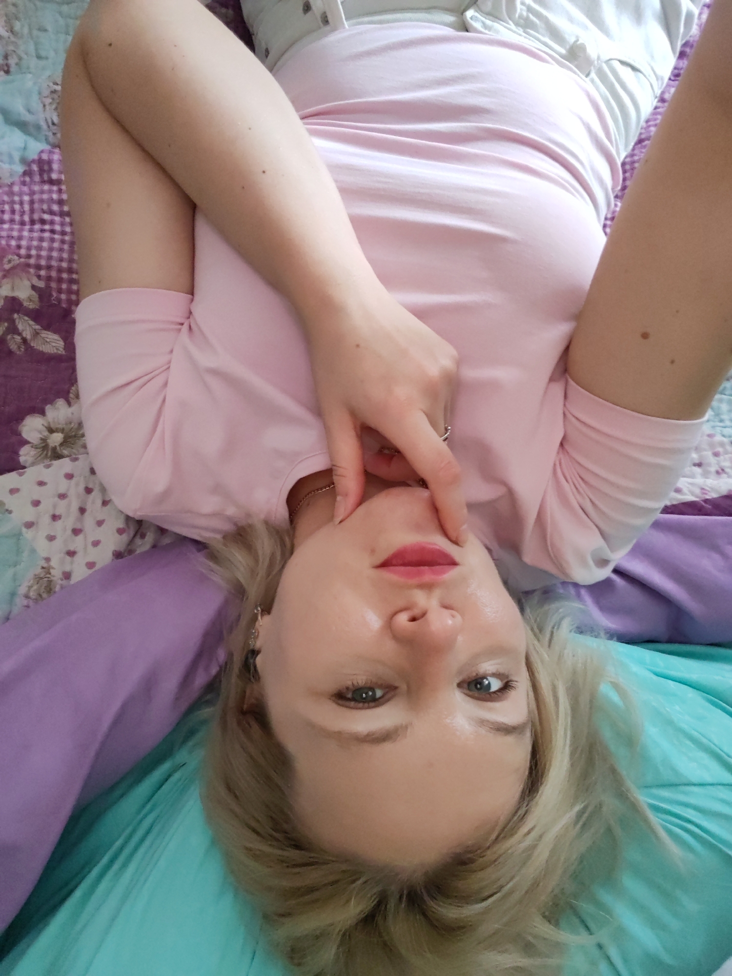 A close-up, upside-down selfie shows a young, blonde woman lying on a bed. She is wearing a pink t-shirt and white pants. Her arms are crossed in front of her chest, and one hand is touching her chin. Her lips are painted a bright pink, and she has light makeup on her eyes. The bedspread is a colorful patchwork of purple, pink, and teal fabrics. The woman appears to be relaxed and playful. The scene is indoors, likely in a bedroom, and the lighting suggests it could be daytime. There is no visible text in the image. The location context is Izhevsk, Russia.