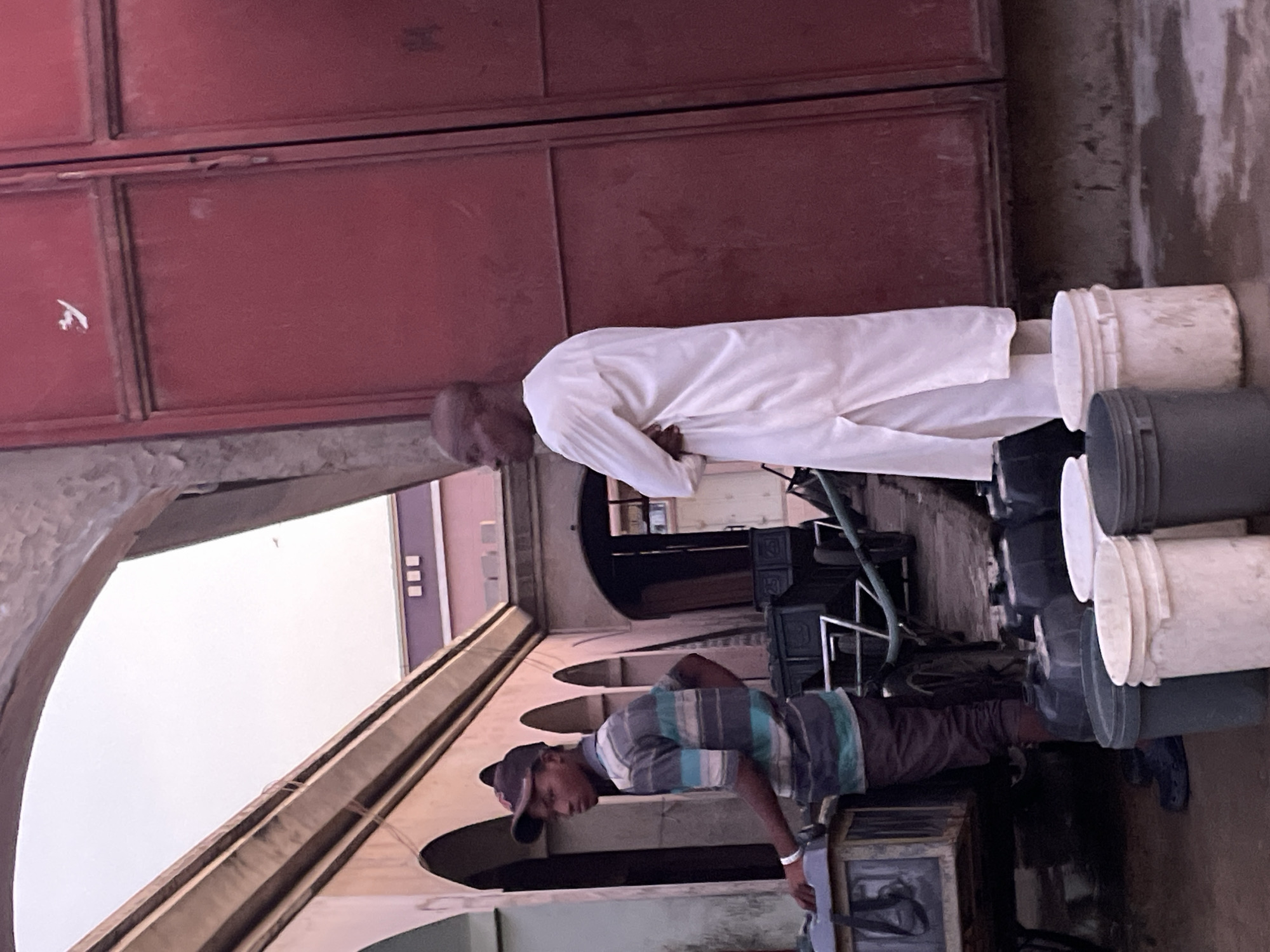 The image depicts a scene likely related to water collection or distribution, featuring two men and numerous buckets set against a building backdrop. The original photo appears rotated 90 degrees clockwise.
In the middle ground, an older man, possibly middle-aged or elderly, stands attentively. He is dressed in a long, flowing white garment, possibly a thobe or similar traditional attire, and has a bald head. He is looking downwards with his hands clasped in front of him, appearing to be observing or waiting.
In the foreground, a younger man is actively engaged in a task. He wears a striped short-sleeved shirt (featuring horizontal stripes of blue, white, and a muted green/grey), dark pants, and a dark baseball cap. He is bent over a structure or object, suggesting he is operating equipment or handling items at a lower level, possibly in connection with the buckets.
To the right of the older man and behind the younger man, several plastic buckets are stacked. These buckets are in varying states of cleanliness and color, with some being white and others dark grey or black. A green hose or pipe is visible near the stacked buckets, indicating a potential water source or delivery system.
The background consists of large, reddish-brown or maroon metal doors, which might belong to a storage facility, workshop, or building entrance. To the left, architectural elements like archways and building facades are visible, with soft, diffused light suggesting an open-air but shaded environment, or possibly an overcast day. No discernible text is visible on any surfaces or objects in the image. The general atmosphere suggests a functional, possibly working, area.