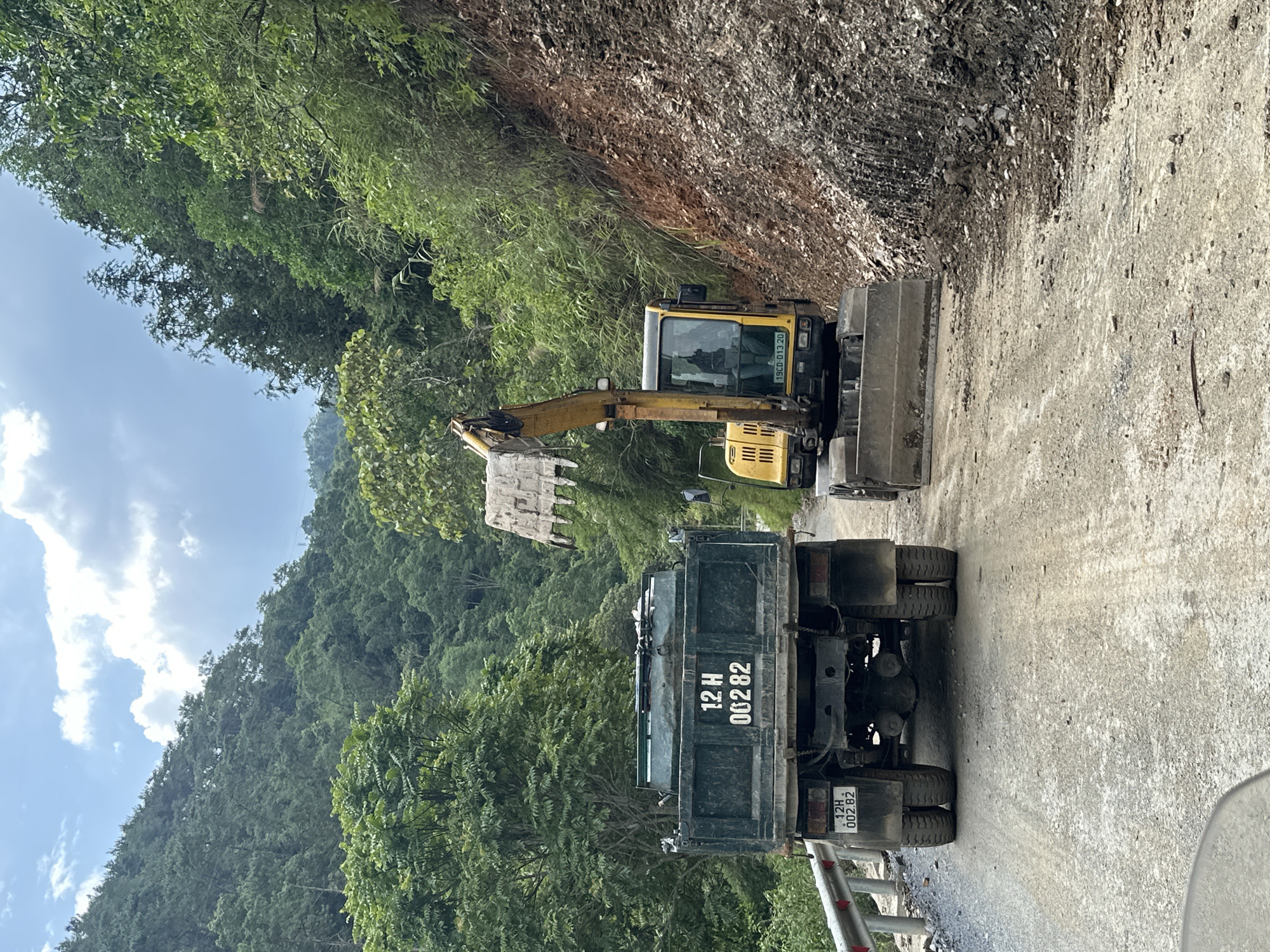 The image shows construction in the Bạch Đích Commune, Vietnam. A yellow excavator is loading dirt into the back of a green dump truck on a narrow road. The area is mountainous with dense green vegetation.