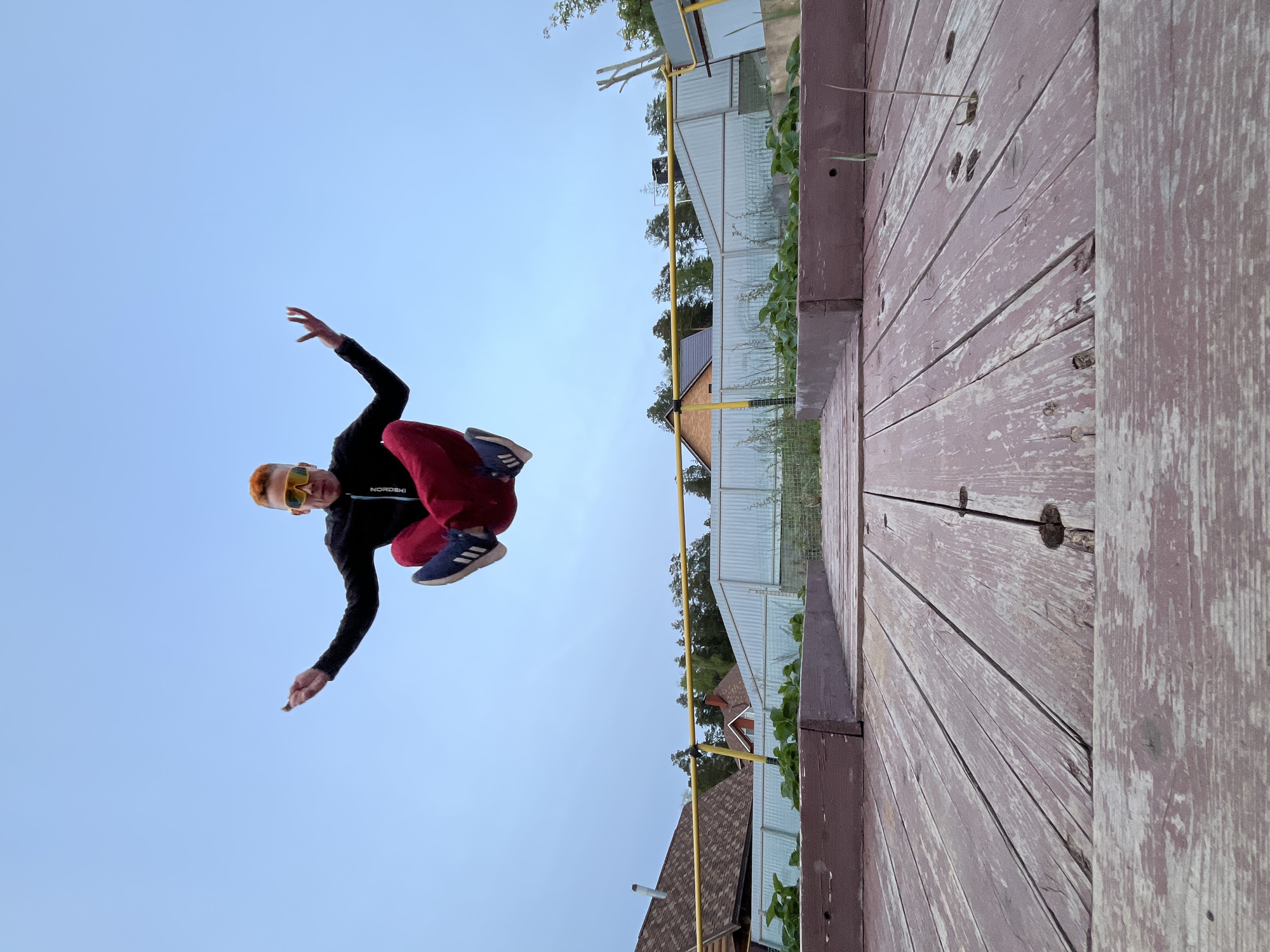 A medium shot shows a man in mid-air, seemingly jumping from a wooden structure. The man is dressed in a black jacket, red pants, and blue sneakers. He is wearing yellow sunglasses and has reddish hair. He is in a dynamic pose with arms and legs outstretched. The background includes a pale blue sky, a wooden structure with planks, and residential buildings behind a fence with green plants. The scene is located in Chaikovsky, Russia.