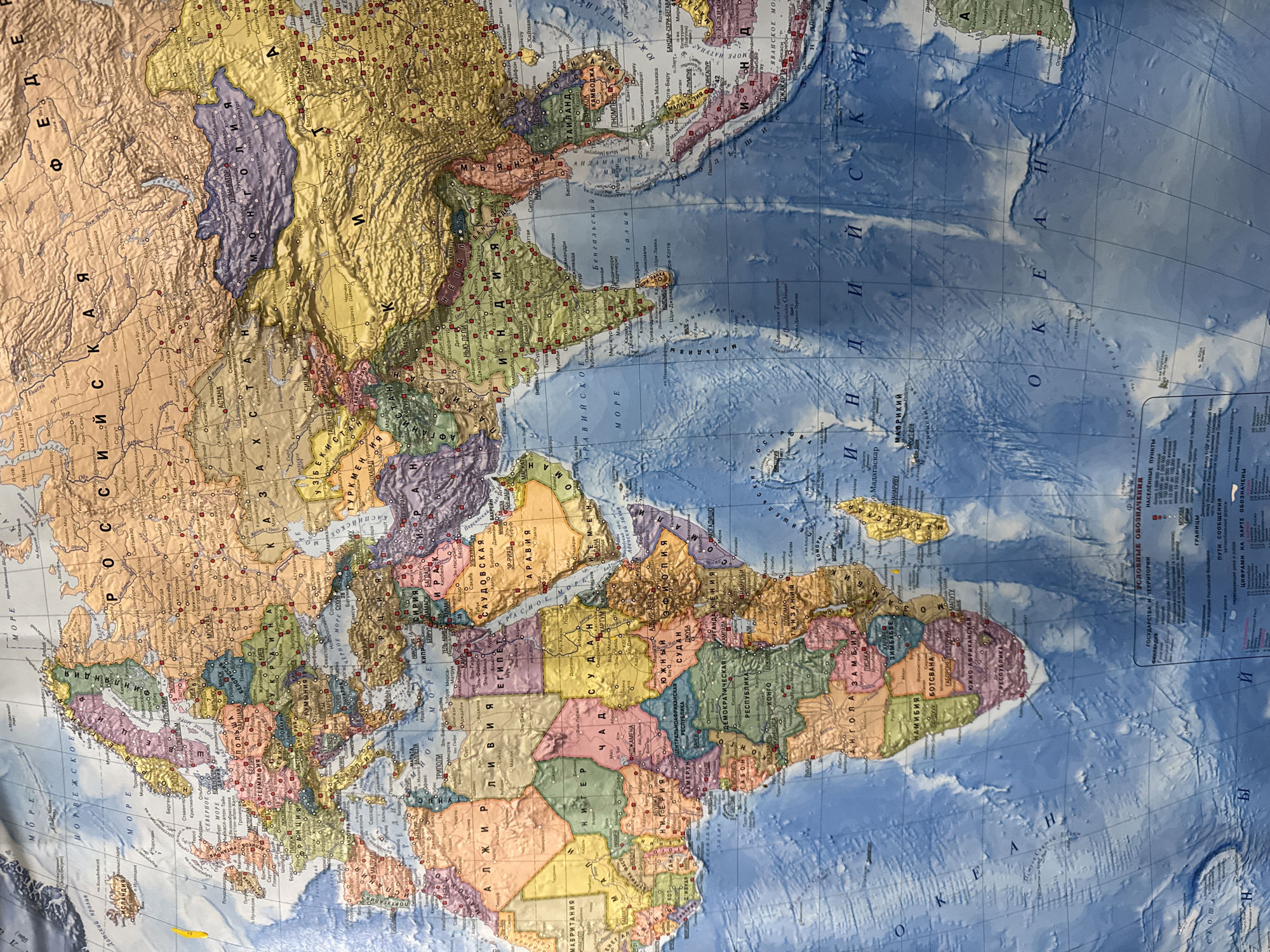 The image is a close-up, angled, full-frame photograph of a world map printed on a curved surface, likely a globe or a large map display. The map is richly detailed, showing countries, cities, bodies of water, and geographical features. The colors are vibrant, with blues for water, various greens, yellows, and browns for landmasses, and distinct colors for political borders. Russian text is prominently visible throughout the map, indicating it is a Russian-language map.
The map displays large portions of Europe, Asia, and Africa, as well as parts of the Middle East and the Indian Ocean. Several cities are labeled in Russian, including Moscow and St. Petersburg in Russia, as well as other international capitals. The map also shows the Ural Mountains and the vast expanse of Siberia.
The location "Sovetskiy, Russia" is not directly visible or identifiable within the cropped section of the map. While the map clearly depicts Russia, specific smaller towns or cities like Sovetskiy are not legible at this magnification and detail level. The focus of the image is on the overall representation of continents and major geographical and political divisions.