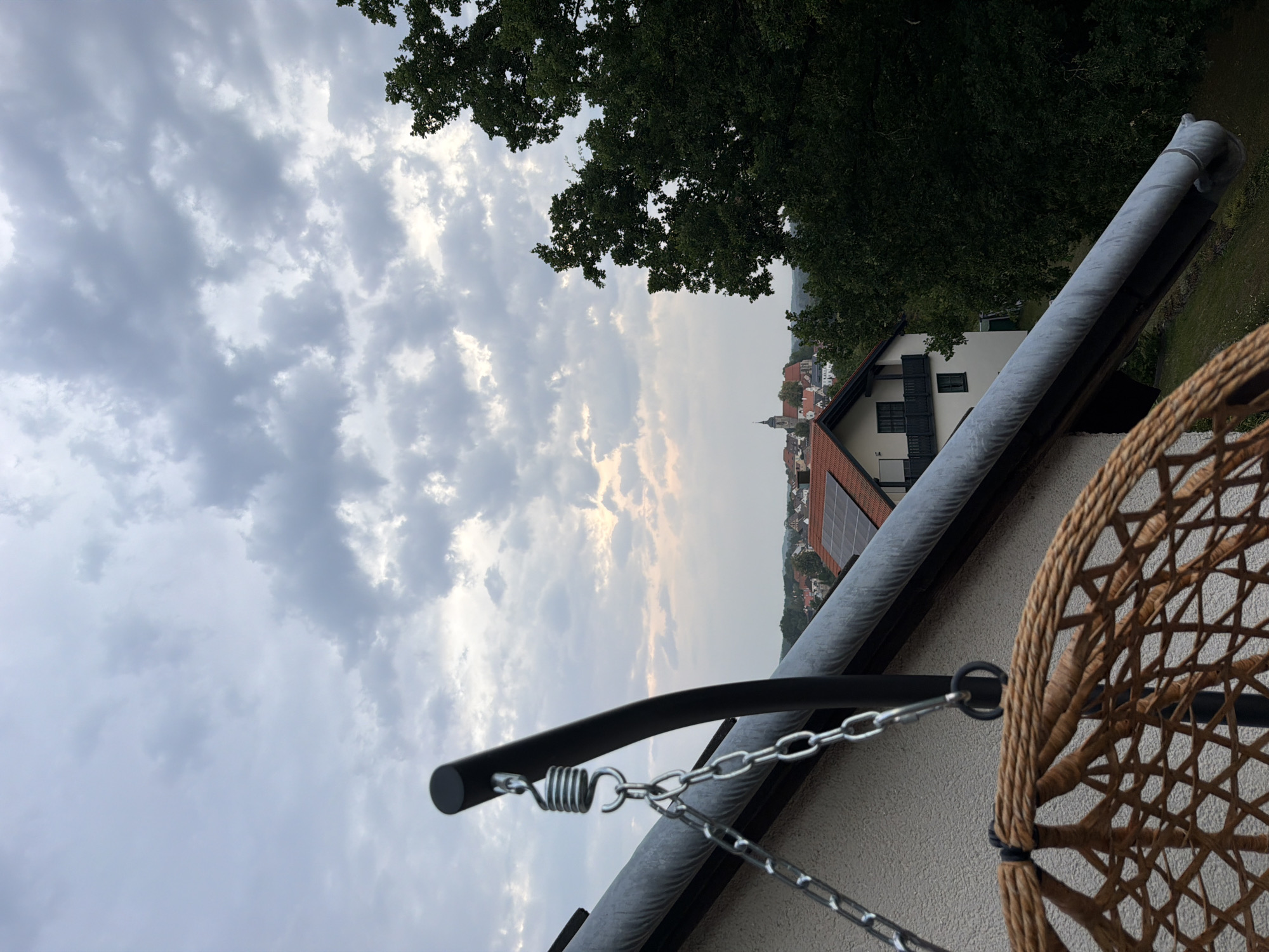 The image captures a view from a balcony or elevated structure overlooking the town of Creußen, Germany. In the foreground, the textured wicker of a hanging chair is visible, along with its supporting metal frame, spring, and chains. A grey downspout runs along the angled roofline of a white house.
Beyond the immediate foreground, a picturesque townscape unfolds under a cloudy sky. Buildings with red-tiled roofs, including a prominent church with a steeple, are clustered together. Solar panels are visible on one of the rooftops. Dense green trees frame the right side of the scene, suggesting a verdant environment surrounding the town. The overall atmosphere is subdued, likely due to the overcast weather. The image appears to have been taken during the late afternoon or early evening.