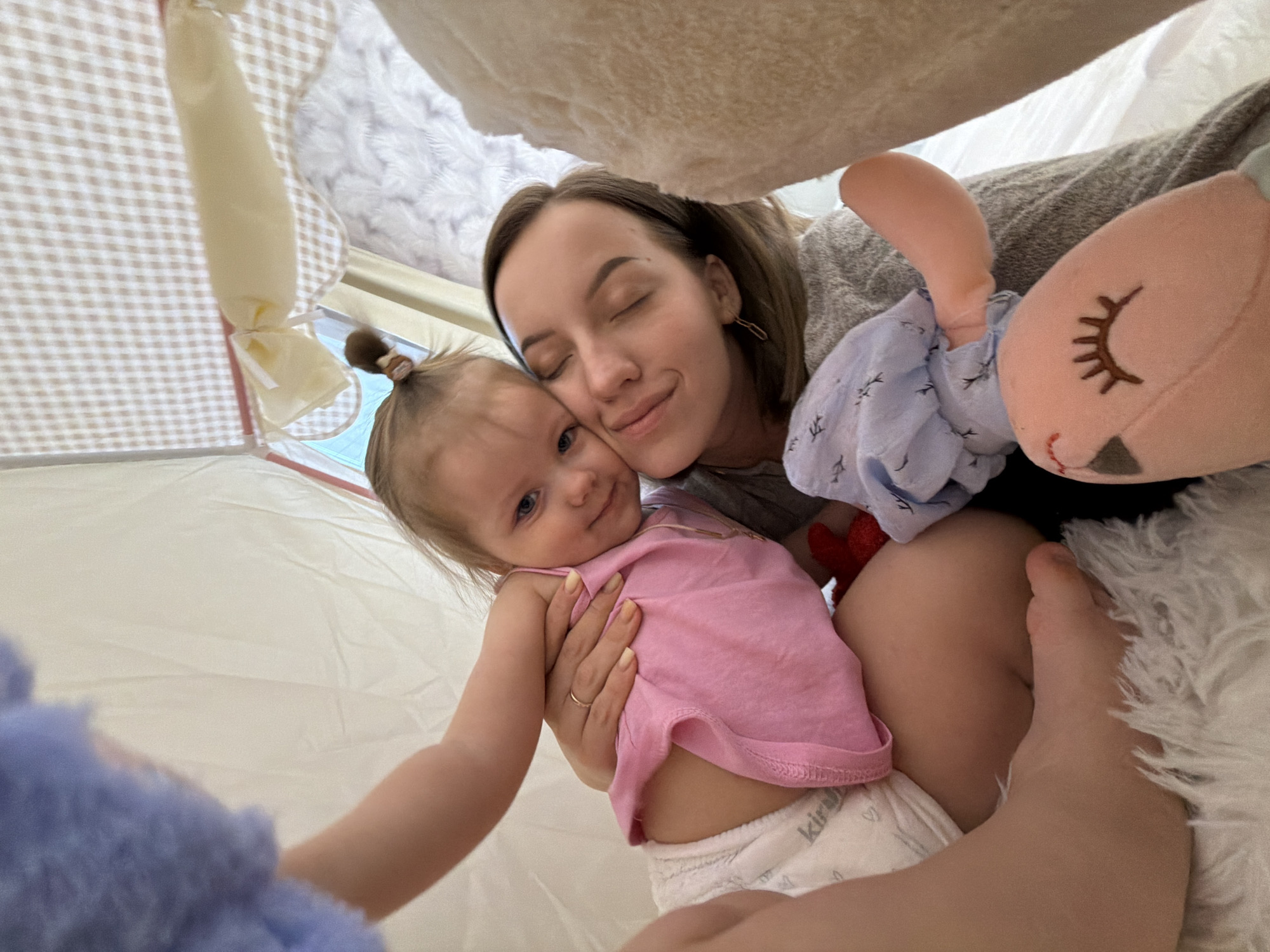 A woman with short brown hair and closed eyes is holding a baby with blue eyes, a pink shirt, and a diaper inside a fabric tent. The woman is wearing a grey shirt and a gold ring. A plush doll with a closed eye and a blue patterned dress is also present. The environment appears to be a domestic setting. The city of Reshetnikovo, Russia is not identifiable in the image.