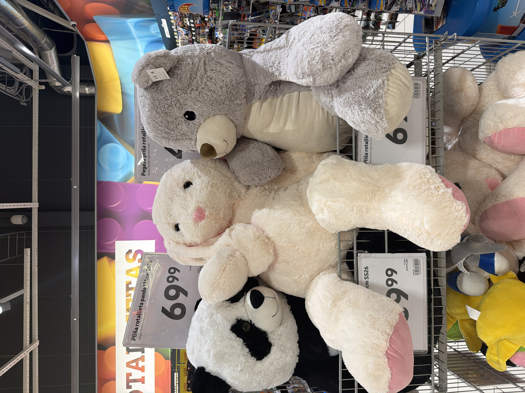 The image captures a detailed view of a retail store's toy section in Krustkalni, Latvia, specifically featuring a display of three large plush animals. In the foreground, from left to right, are a black and white panda, a cream-colored bunny, and a light grey bear, all large in scale and positioned upright within a metal display cage or shelf. The panda is on the left, looking straight ahead, with the bunny situated to its right, resting its left arm across the panda's side. The bunny features floppy ears with pink inner lining and a pink stitched nose. To the bunny's right is the grey bear, with a brown snout and a cream-colored belly, its left arm draped over the bunny. A small white tag displaying a "CE" mark is attached to the bear's left ear.
In front of the panda and bunny, a white rectangular price sign, encased in clear plastic, displays text in Latvian: "Plīša rotaļlieta panda 136cm SS26," indicating a 136cm panda plush toy. The prominent price "69⁹⁹" is printed in large black numerals. Below this, smaller text reads "Jiangsu Runshi Light and Textil," along with a barcode and a secondary price of "69,99 €/gab." Another partial price sign is visible to the right, showing "Pegaza plīša rotallieta" (Pegasus plush toy) and a partially obscured price beginning with "6." and also "69,99 €/gab."
Below the main plush toys, on a lower shelf of the metal display, various other smaller plush toys are visible, including several in yellow and some with blue and white accents. A blue plastic bin is visible to the far right. The background features colorful wall murals with large, abstract shapes in yellow, blue, purple, and orange. Above, the store's ceiling is dark grey with visible industrial-style lighting fixtures, exposed ventilation ducts, and metal grating. Other retail shelving stocked with merchandise, including packaged "Hot Wheels" toy cars, is visible further back on the right side of the frame. Latvian signs such as "ROTAĻLIETAS" (TOYS) and "AUTOMAŠĪNAS" (CARS) are discernible on the distant shelving units. No individuals are present in the image.