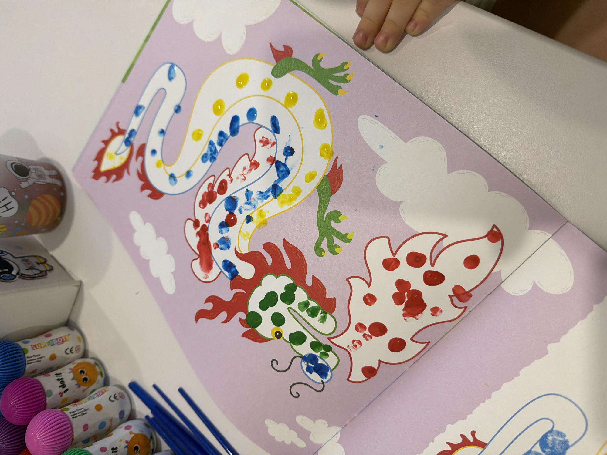 A close-up shot shows an open book on a light-colored surface. The book features illustrations of Chinese dragons outlined in red, with colored dots in red, blue, green, and yellow used to fill in the shapes. One dragon's body is white, while its head is white with green dots. The background of the book page is a light purple color, with drawings of white clouds. To the left of the book are several paint daubers with colored pom-poms and blue paint brushes. A small can featuring an illustration of an astronaut, and a white box are also visible. The edge of a hand appears in the upper-right corner of the frame. The location is unspecified, but given the request to include Chaikovsky, Russia if available, the image was likely taken in that city.