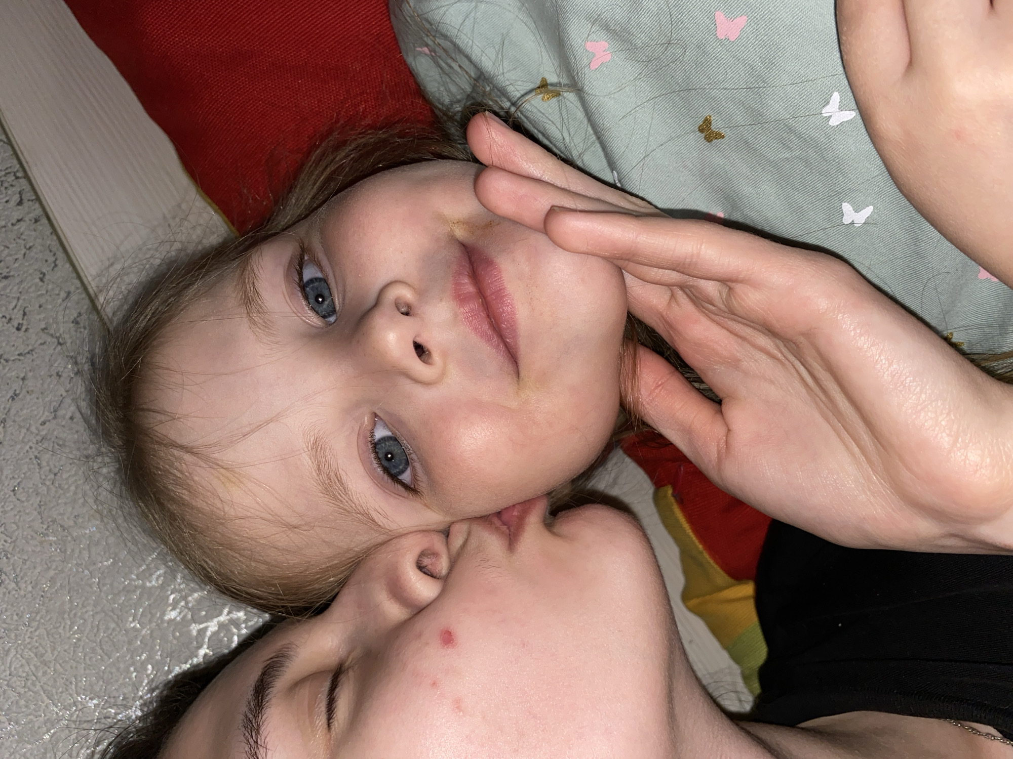 This intimate portrait, taken in Votkinsk, Russia, captures a close moment between an adult and a young child, likely a parent and their offspring. The subjects are positioned lying down, with the child's head in the upper portion of the frame and the adult's head below, nuzzling or kissing the child's cheek/nose area.
The child, appearing to be a young girl, has fair skin, light blonde or brown hair, and striking blue eyes that gaze towards the viewer. Her lips are slightly parted. One hand, presumably the adult's, gently rests on the child's face near her mouth. She is partially covered by a light green fabric adorned with small pink and gold butterfly patterns. Behind her head, a textured red pillow or blanket is visible.
The adult's face is mostly obscured by the child's head, but her mouth, closed in an expression of affection, is visible against the child's skin. A small red blemish is noticeable on the adult's cheek. Part of a black garment and a silver chain are visible on the adult's body.
The setting appears to be indoors, likely a home, with a light-colored, textured surface (possibly a floor or wall) forming part of the background. The lighting is soft, suggesting an intimate and calm atmosphere. The overall emotion conveyed is one of tenderness, love, and familial bond, captured in a quiet moment. No visible text is present.