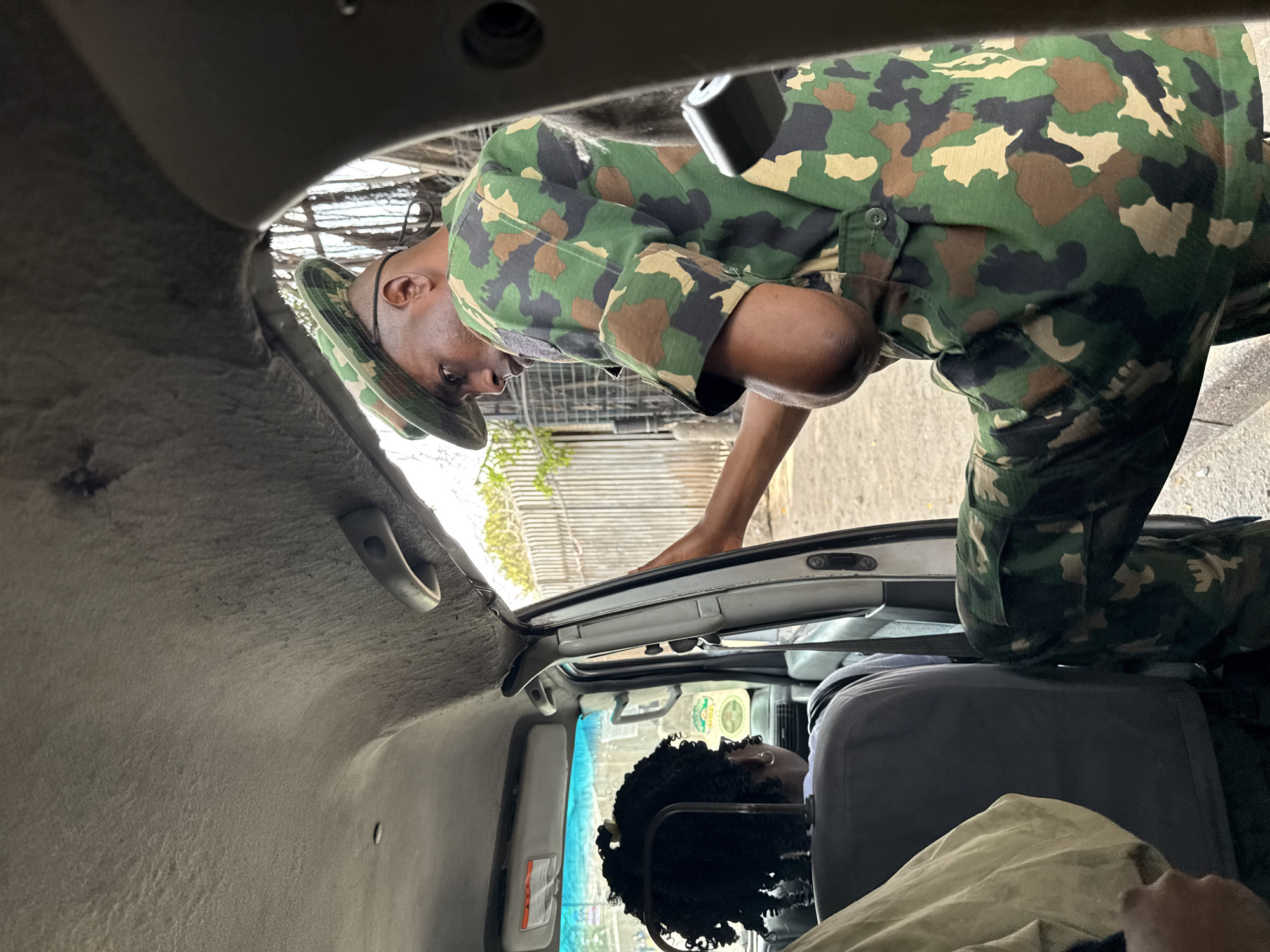 The image shows the interior of a vehicle, likely in Lagos, Nigeria, based on context. A person in military camouflage uniform is leaning into the vehicle, presumably interacting with the passengers. The passengers are visible seated in the back. The interior of the vehicle is functional and shows signs of use. The outside environment is partially visible through the vehicle's open rear and side windows.