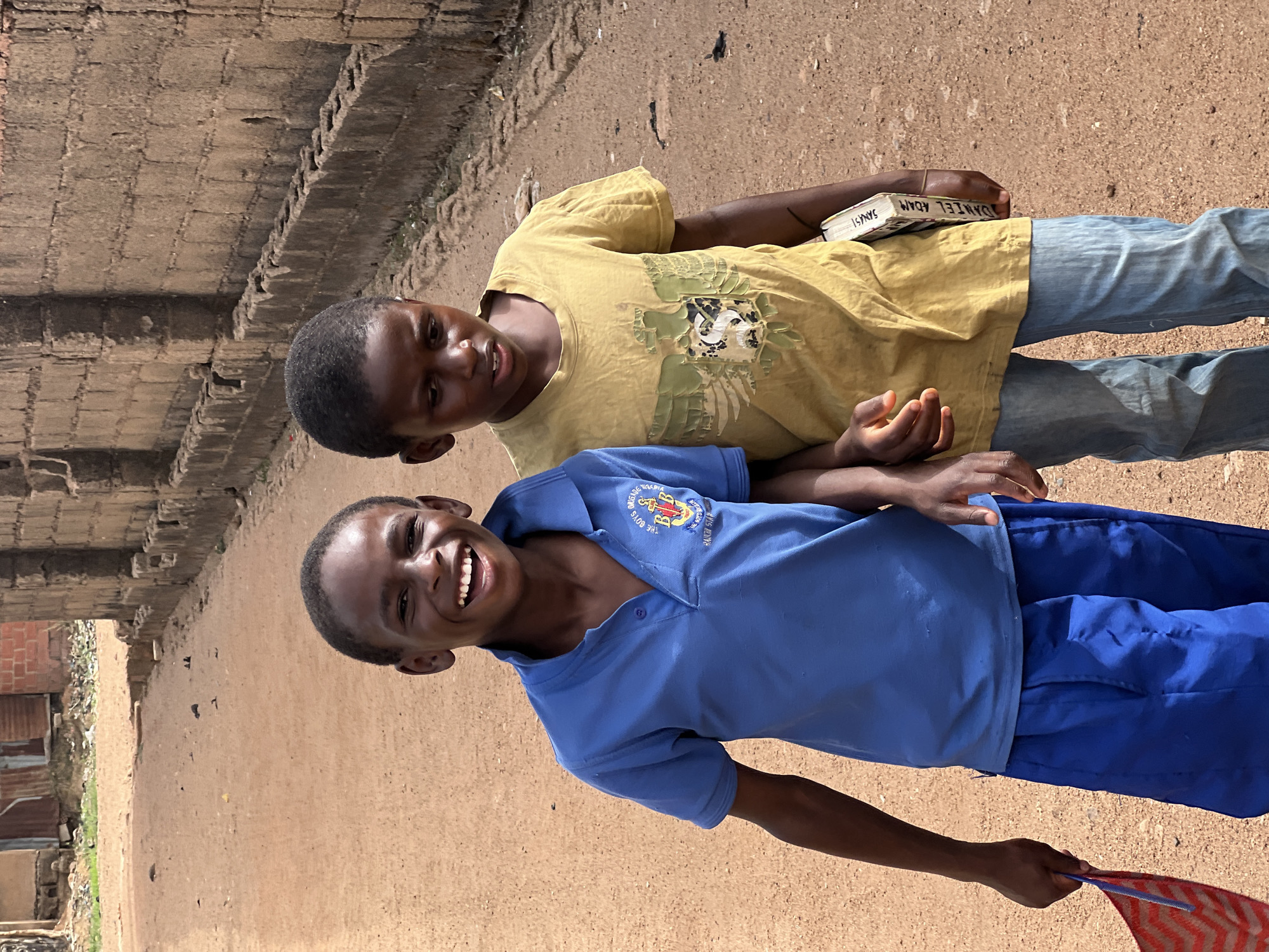Two young boys, likely children, are positioned outdoors in what appears to be Bukuru, Nigeria. The boy in the foreground is wearing a blue polo shirt, adorned with a crest emblem, and blue pants, indicating a school uniform. He is smiling broadly, revealing his teeth. The boy in the background is wearing a yellow t-shirt with a camouflage-like pattern and blue jeans. He is holding a book labeled "Daniel Adam". The ground is a dusty, reddish-brown dirt. Behind them are concrete structures, and in the distance are buildings with corrugated metal roofs. The photo shows signs of poverty and a less-developed environment.
