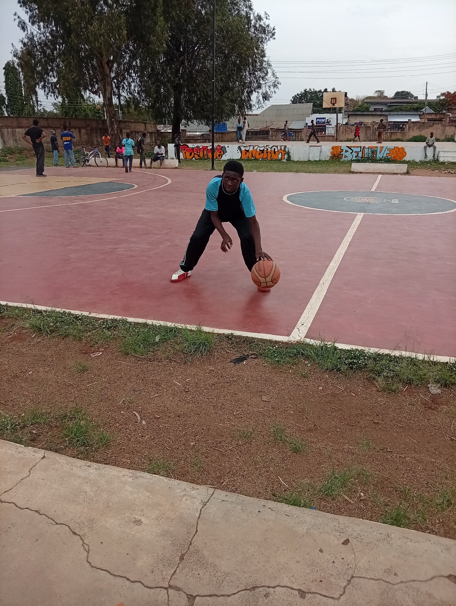 On an outdoor basketball court in Unknown, Unknown, a young man dribbles a basketball. He wears a black and blue shirt and black pants. The court is red with white lines and a blue circle. Other people are visible in the background, some standing, some sitting, and one riding a bicycle. A low wall behind the court is covered with graffiti. The sky is overcast. A tree stands to the left of the court, with buildings visible in the distance. The foreground is a dirt and grass area with a concrete curb.