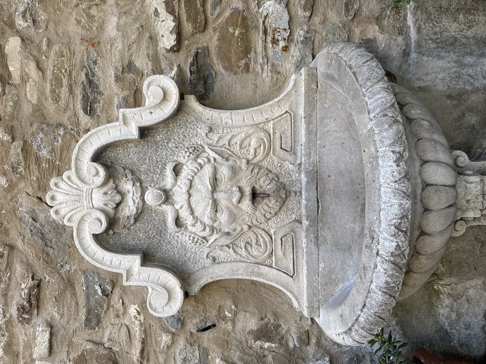 The image shows a weathered stone fountain mounted on a stone wall. The fountain features a carved human-like face with a beard, horns, and grapes in its hair, from whose mouth a small spout emerges. Below the face is a basin with a decorative rim. The wall behind the fountain is constructed from irregular stones of various sizes and colors, some appearing rough and others smoother. The overall aesthetic suggests an older, rustic setting, consistent with the historic architecture found in towns like Dolceacqua, Italy. There are no people or visible actions in the image. A few green plants are present to the left and right of the fountain, suggesting it might be in an outdoor or garden area.