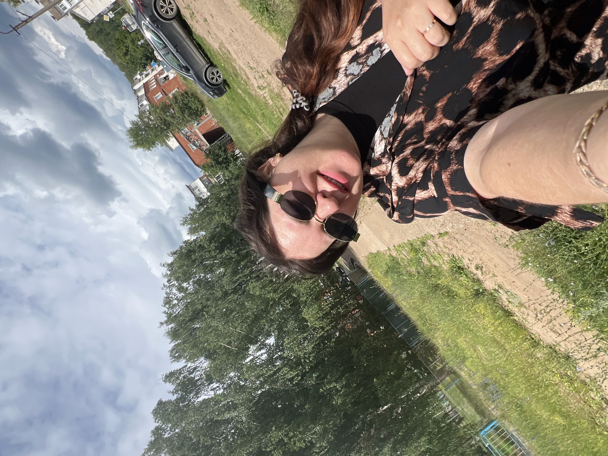A person, likely a woman, is taking a selfie outdoors on a sunny day. She is wearing a black top with a leopard print dress over it, sunglasses, and a ring on her finger. Her hair is dark brown and pulled back in a ponytail with a scrunchie. She is smiling at the camera. In the background, a dark-colored car is parked on a dirt road, with some buildings and trees visible. The weather appears to be clear and sunny, with some clouds in the sky. The overall scene suggests a casual, relaxed atmosphere. There is no visible text in the image.
