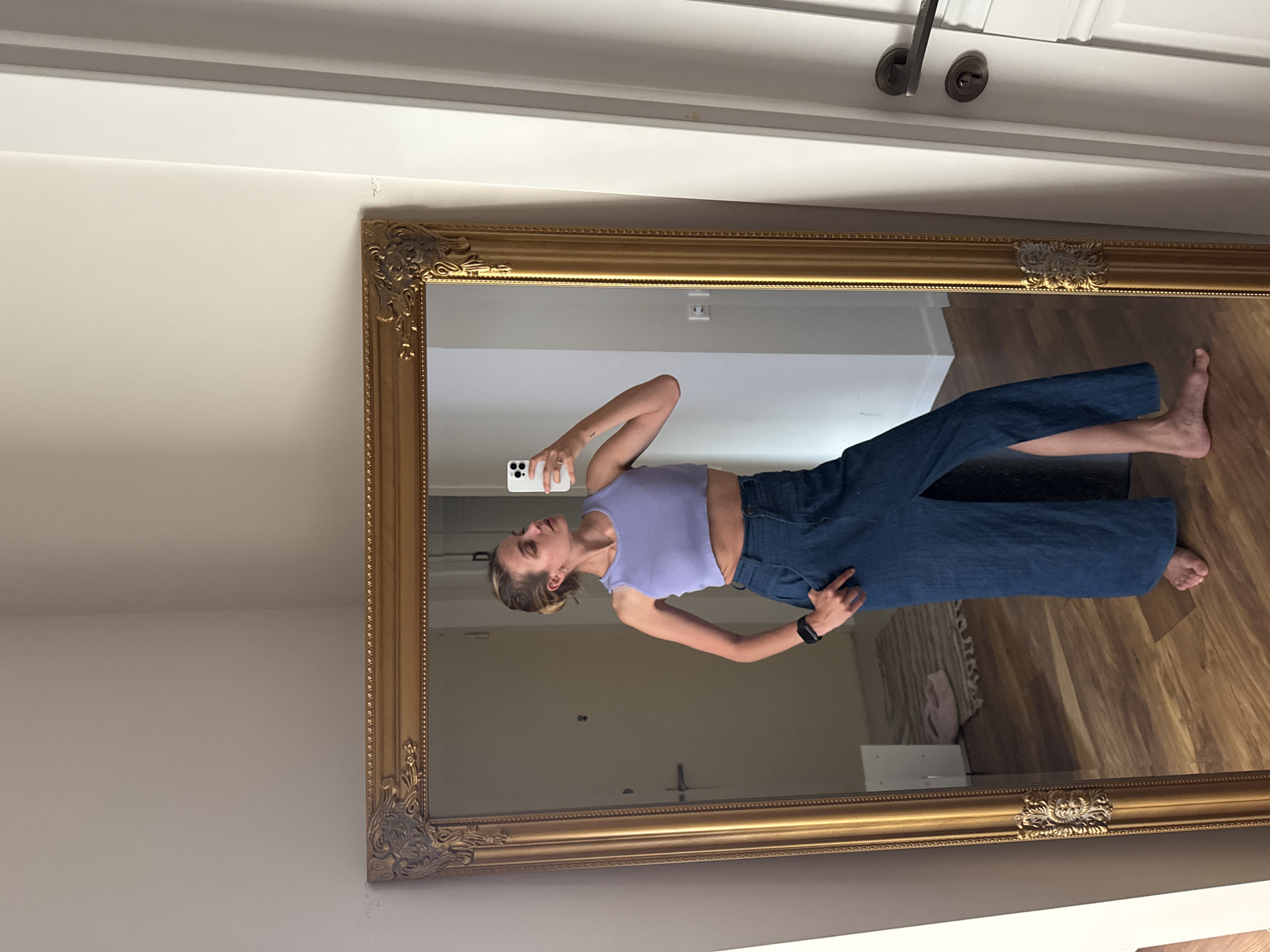 A woman is seen in the reflection of a large ornate gold-framed mirror. She is holding a smartphone, presumably taking a photograph of herself. She is wearing a light purple top and dark blue jeans. Her hair is pulled back in a bun. The mirror is mounted on a white wall with a white door frame visible at the top. The reflection shows a room with wooden flooring.