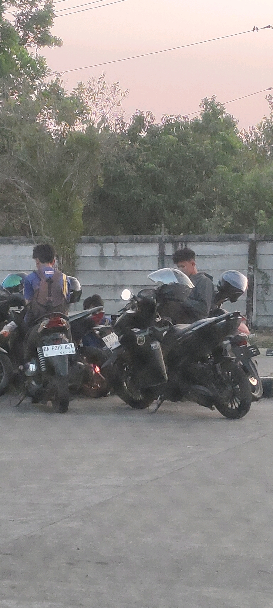 The image depicts a street scene in Bentok Kampung, Indonesia, with several parked motorcycles and a few individuals. The foreground is dominated by the asphalt pavement. In the mid-ground, multiple motorcycles are parked, some with riders. One motorcycle in the foreground has a license plate that reads "DA 6213 BCX". A person wearing a blue shirt and a backpack is visible near the motorcycles. Behind the motorcycles, a grey concrete wall runs horizontally across the frame, with trees and foliage above it. The background features more trees and a light-colored sky, suggesting it might be early morning or late afternoon. Power lines are also visible against the sky.