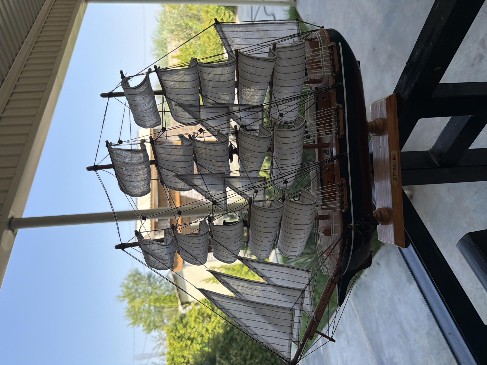 A detailed model of a historic sailing ship, the Cutty Sark, is displayed outdoors. The ship is intricately constructed with numerous white sails, a dark hull, and wooden masts and rigging. It rests on a wooden stand that bears the inscription "CUTTY SARK 1869". The ship is positioned on a black metal railing, suggesting it's on a balcony or patio. Behind the ship, lush green trees and foliage are visible, along with hints of buildings. The sky is clear and blue. The lighting indicates it is daytime, likely sunny. The image appears to be taken in a residential or public outdoor space. The context provided about Sabirabad, Azerbaijan, places this scene within that specific geographical location.
