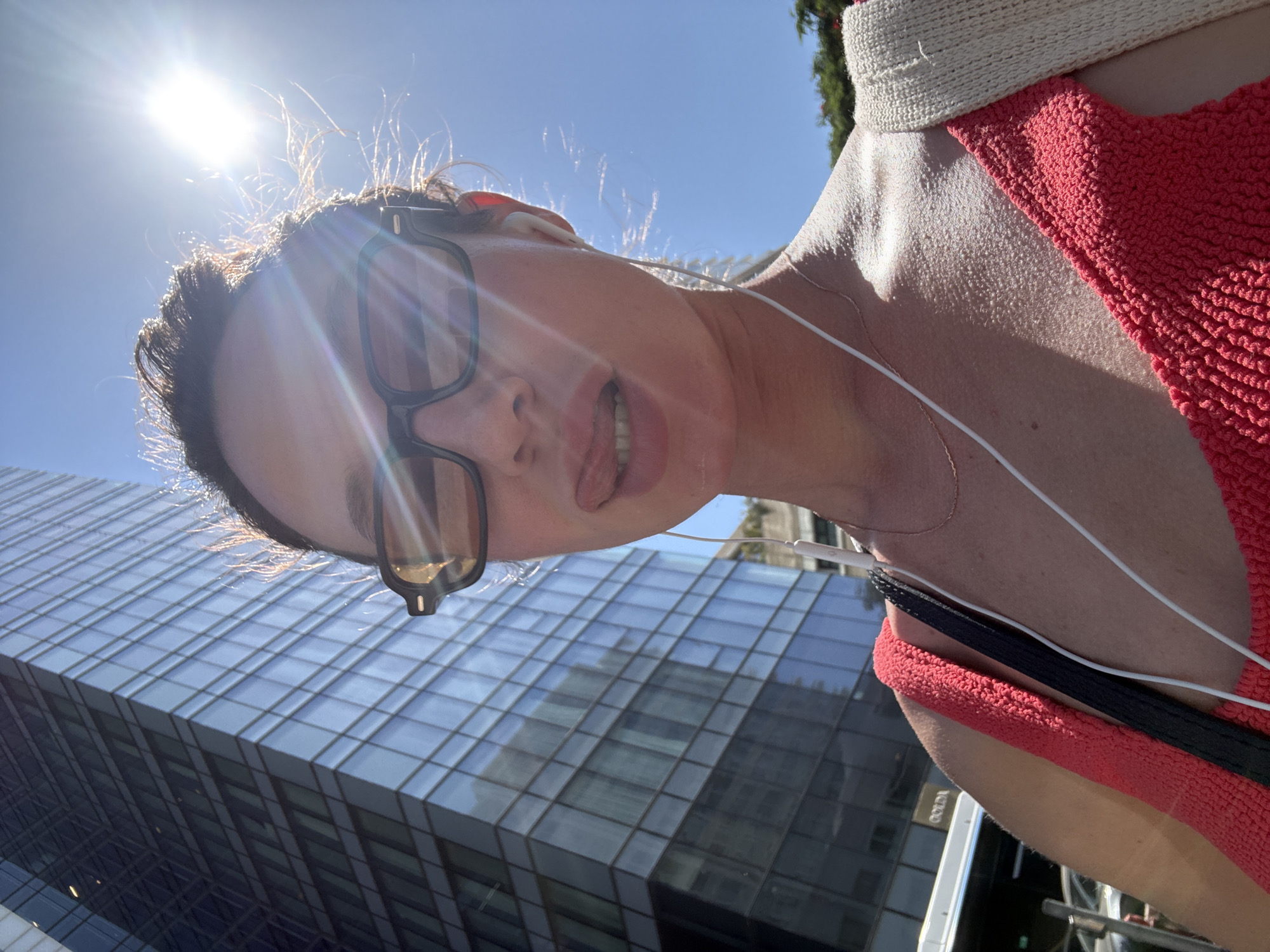 A woman is captured in a close-up selfie, looking directly at the camera with her tongue slightly extended. She wears dark-framed sunglasses with amber-tinted lenses, through which strong sun glare is visible. White wired earbuds are in her ears, with the wires extending down her chest. Her dark hair is pulled back, with individual strands backlit by the sun, creating a halo effect. She is wearing a coral-red, textured sleeveless top, and a thin gold-colored necklace is visible. A black strap crosses her left shoulder, and a beige woven strap is visible on her right shoulder. The bright sun is intensely visible in the upper right portion of the frame against a clear blue sky. In the background, a large, modern glass skyscraper with multiple reflective windows dominates the left side of the image. At the lower left, below the building, a white vehicle, possibly a bus, with the partially visible word "GOLDA" on its side, is parked. Green foliage is faintly visible in the far distance on the right. This scene is located in the city of Tel-Aviv, Israel, as indicated by the "GOLDA" branding, which is associated with a prominent Israeli ice cream chain.