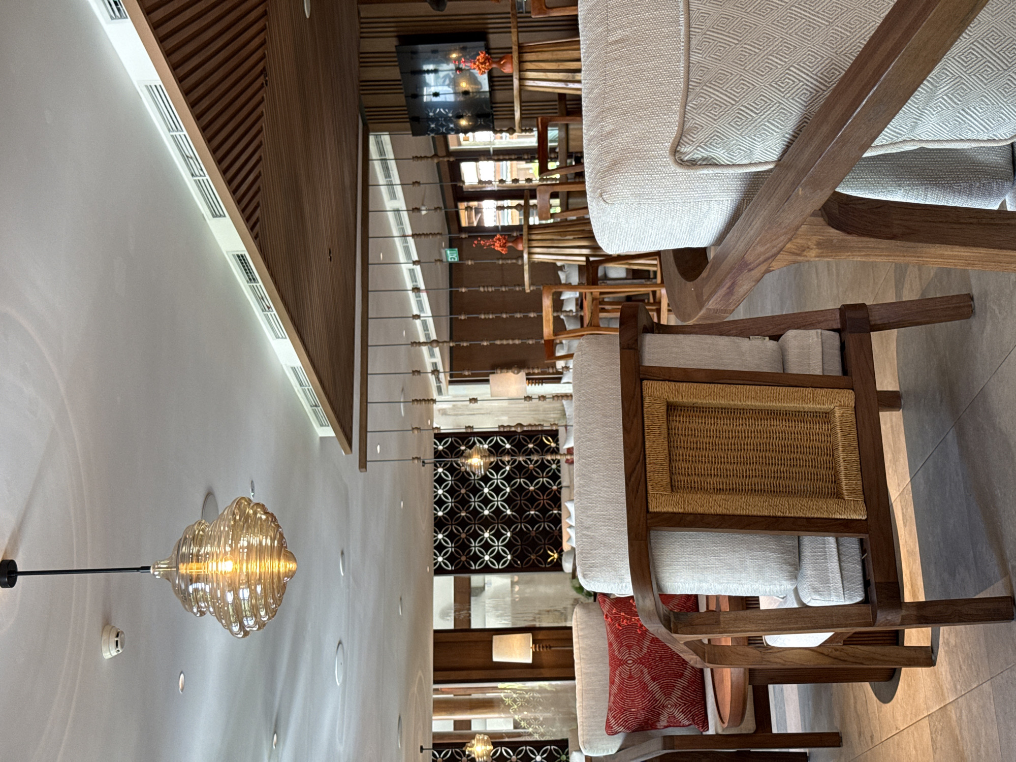 The image depicts a well-appointed indoor lounge or waiting area, likely within a hotel or resort, in Kuta Selatan, Indonesia. No people are visible, suggesting a tranquil atmosphere.
The primary subjects are comfortable wooden armchairs with light-colored, patterned cushions. One chair prominently features a woven rattan backrest, and another has a decorative red patterned throw pillow. The floor is covered in large, light-colored tiles.
Architectural details abound, including a striking wooden slatted ceiling or wall section. A long, white ceiling panel runs along the top-left, fitted with several linear air vents. A visually interesting decorative glass pendant light, with a warm glow and a ribbed or fluted design, hangs from the ceiling, alongside multiple recessed spotlights and a smoke detector. In the background, a large decorative metal screen with an intricate geometric, interlocking circle pattern acts as a room divider or architectural feature. Further back, a dark rectangular screen or artwork is mounted on a wall, with two small orange vases placed below it.
An "EXIT" sign with green lettering and an arrow pointing left is visible in the mid-ground. The overall ambiance created by the warm lighting, natural materials like wood and rattan, and sophisticated design elements suggests a relaxed and elegant setting, indicative of a tropical resort environment. The time of day appears to be evening or a well-lit interior, relying on artificial light sources for illumination.
