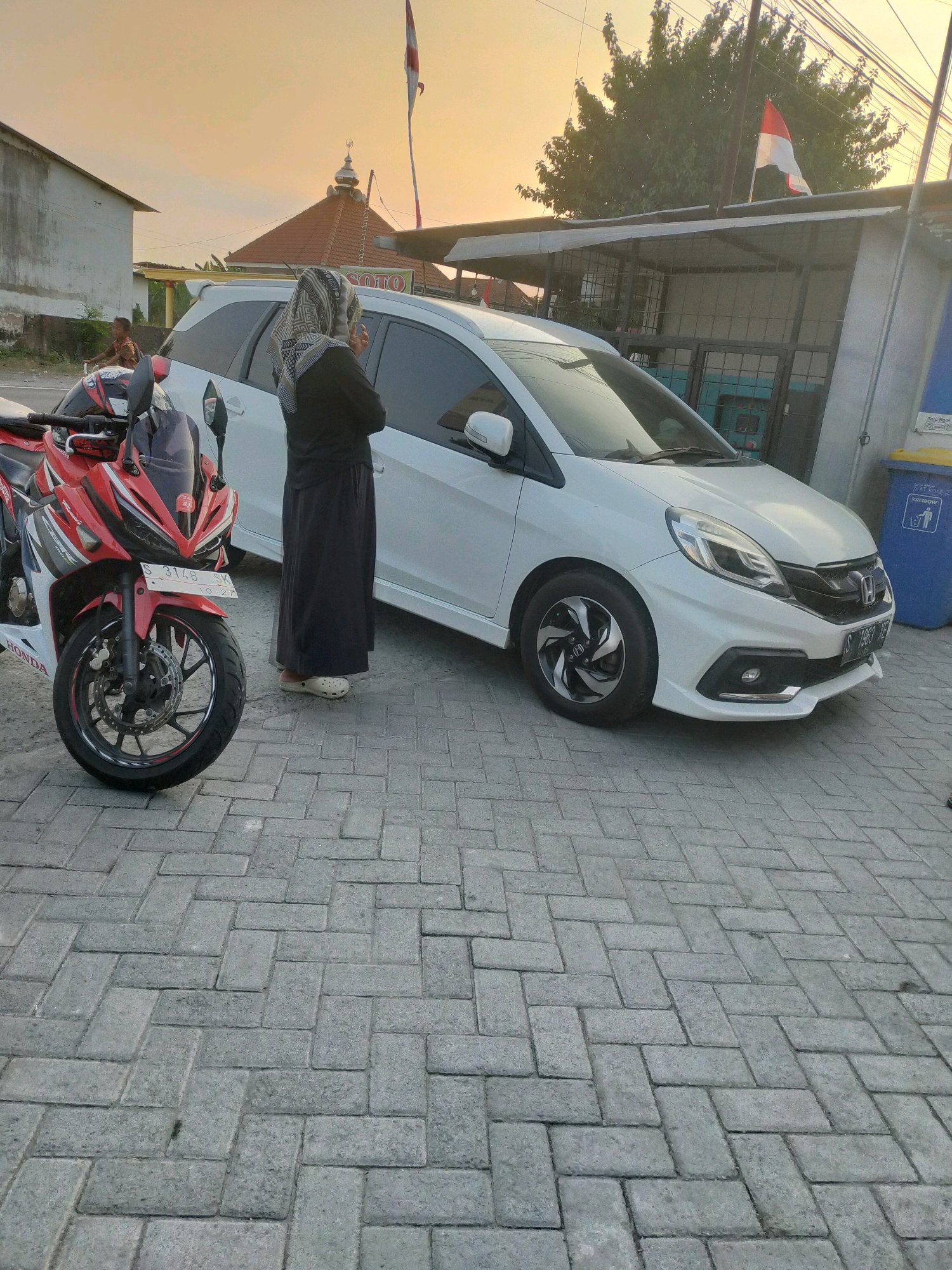 In the city of Puskesmas Keboan, Indonesia, a white car and a red and white motorcycle are parked on a paved stone driveway. A person wearing a black abaya and a patterned hijab stands between the two vehicles, facing away from the camera. In the background, a building with a red-tiled roof and a dome is visible, along with trees and overhead wires. The sky is a warm orange, suggesting it is either sunrise or sunset. A red and white flag is also visible, waving from a pole near the building. The scene appears to be outdoors, likely in a public area or a residential street.