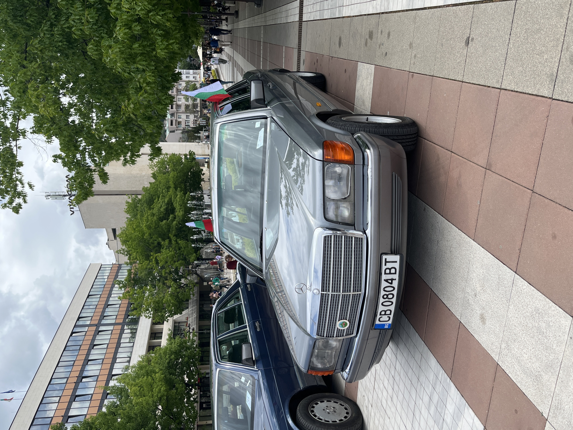 The image shows a gray Mercedes-Benz car parked on a sidewalk in an urban area. The license plate reads "CB 0804 BT". The car is angled towards the right of the frame, and a dark blue car is partially visible to its left. The background includes a tall building with multiple floors, trees, a public square with a fountain, and people gathered in the distance. Bulgarian flags are present, suggesting a possible celebration or event. The sidewalk is paved with a mix of gray and brown tiles. The sky is overcast. The location appears to be a city square in Bulgaria, given the license plate and flags.