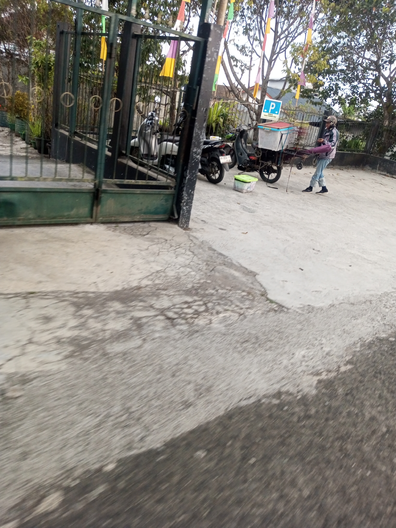 A person is standing near two motorcycles on a paved area in front of a green metal gate. The person, wearing a cap, jacket, and jeans, is holding a purple object and appears to be interacting with a cart attached to one of the motorcycles. The cart has a blue cooler on top. A blue and white parking sign is visible on a pole behind the cart. The environment suggests an outdoor, possibly residential or commercial, setting in Pandansari, Indonesia, with trees and flags in the background. The ground is made of concrete with some cracks and patches.