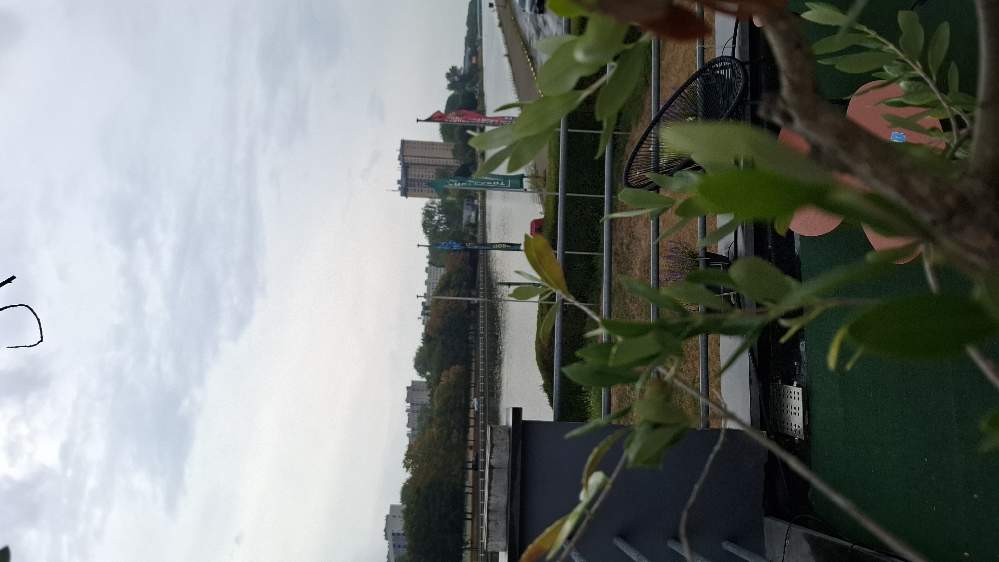 The image depicts a view across a body of water in Ghent, Belgium, likely the canal alongside the Dampoort or bordering the Sint-Pieterspark, looking towards the east. In the foreground, a dark grey railing runs horizontally across the lower portion of the frame. Through a blurry foreground of green foliage, one can see a green carpeted area on what appears to be a balcony or deck. Two light pink, circular objects, possibly furniture or decorative items, are visible on this green surface. Behind the foliage, a black, woven-style chair is situated on the same level. Further into the mid-ground, a serene body of water stretches out, bordered by a paved walkway on the far side. Several tall, slender flags, predominantly blue and green with some red, are planted along this walkway, some featuring white lettering. Beyond the water and walkway, a line of deciduous trees with autumn foliage forms a verdant border. In the background, a cluster of apartment buildings and a prominent, tall, light-brown residential tower are visible, characteristic of urban architecture. The sky is overcast with a pale, diffused light. A thin, dark, wire-like object is faintly visible in the upper portion of the sky, disconnected from any discernible structure.