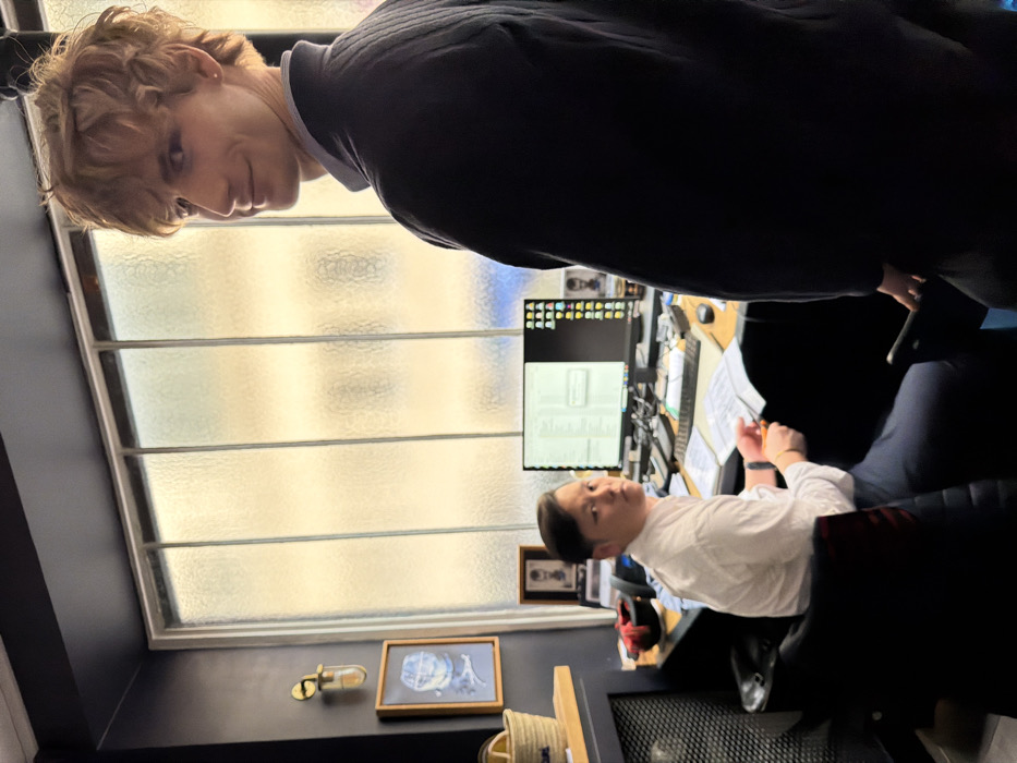 The image depicts an interior office scene in Paris, France. In the foreground, positioned to the right, a fair-skinned male with light brown, wavy hair, wearing a dark quilted jacket over a light blue collared shirt, is seen looking over his right shoulder towards the viewer with a slight smile. He holds a dark rectangular object, possibly a mobile device, in his left hand.
In the background, to the left, an East Asian male with dark, neatly combed hair, wearing a white collared shirt and dark trousers, is seated in a black office chair at a wooden desk. He is holding a pair of scissors in his right hand and has turned to look towards the viewer with a neutral, slightly questioning expression. The desk is cluttered with office equipment, including a computer monitor displaying a split screen with documents on the left and desktop icons on the right, a keyboard, a mouse, a telephone, and various papers. Two framed photographs are visible behind the desk, with one partially obscured by the monitor.
The environment includes a dark-colored wall on the left, featuring a brass-toned wall sconce with a clear glass enclosure and a framed artwork depicting a blue and white nautical or abstract motif. Below these, a dark cabinet or counter supports a wicker basket. The background is dominated by a large window composed of multiple vertical frosted or textured glass panes, obscuring the exterior view but admitting light. A dark grille, possibly covering a radiator, is visible at the lower left.