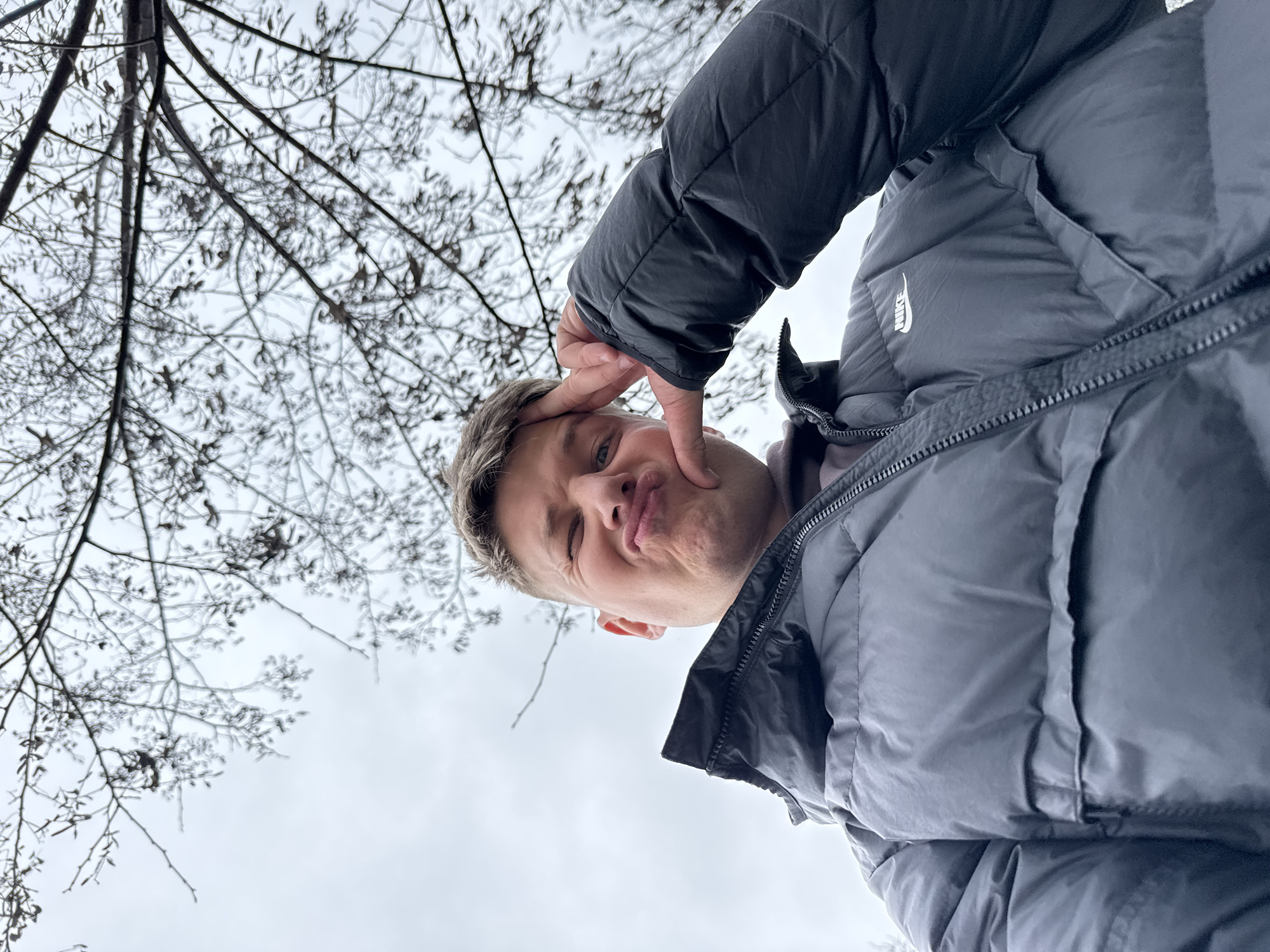 A low-angle, upward-tilted view captures a person wearing a dark puffer jacket with a Nike logo, posing with a playful expression. The person is touching their face with their fingers, making a face with pursed lips and winking one eye. Behind the person, a network of bare tree branches extends across the pale, overcast sky. The location is not evident from this image alone. It would not be possible to locate this image in the city of Plovdiv, Bulgaria.