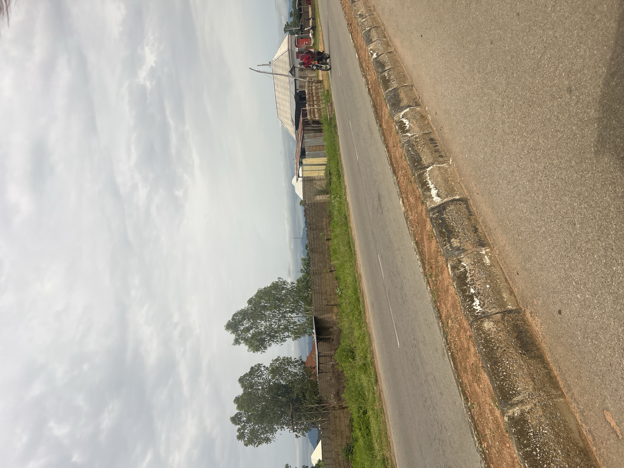 The image depicts a scene on a road in Jalingo, Nigeria, under a cloudy sky. A paved road occupies the foreground and midground, with a worn concrete curb and a dirt shoulder separating it from an adjacent grassy area. Beyond the grass is a concrete block wall, behind which are various structures including corrugated metal sheds, some with visible supporting poles, and buildings with corrugated metal roofs. Two large trees with sparse foliage stand prominently behind the wall. Further in the background, more buildings and distant hills or mountains are visible. A motorcycle with two riders, both wearing red tops, is traveling on the road in the midground, heading away from the camera. The overall impression is of a rural or semi-urban environment.