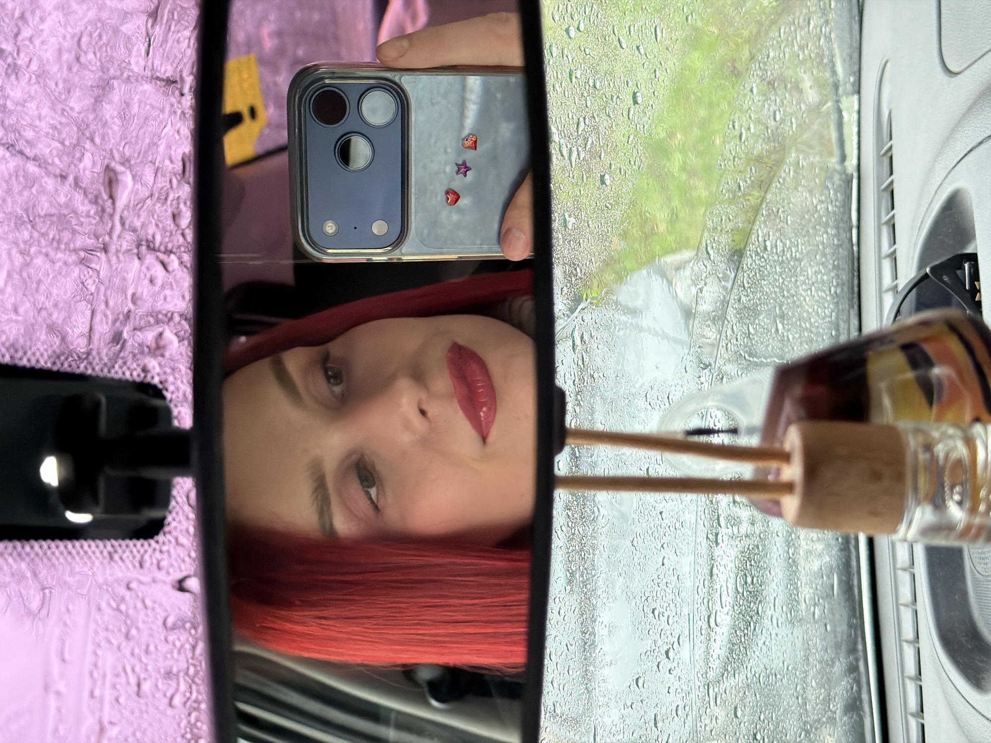 This landscape-oriented image is a selfie taken inside a car, primarily capturing a reflection in the rearview mirror. The central feature is the rearview mirror, presented as a vertical rectangular strip in the middle of the frame.
Reflected within the mirror, and appearing rotated 90 degrees clockwise, is a person's face, likely female. They have vibrant red hair and are wearing red lipstick. Their left eye is visible, gazing towards the right side of the frame. The person's hand is holding a smartphone, also reflected on its side, revealing its rear with a triple camera setup and three small decorative stickers: a red heart, a red star, and an iridescent square.
To the left of the rearview mirror, visible through the car's windshield, is a textured, light purple surface covered in water droplets, possibly the car's headliner or a sun visor. To the right of the mirror, through a rain-streaked window, green foliage (likely trees or grass) is visible, indicating an outdoor scene. The windows are heavily covered in water droplets, signifying active rainfall.
Further to the right of the mirror, parts of the car's interior are visible, including a white plastic air vent and the top section of a car air freshener with wooden reeds, which appears horizontal due to the image's orientation. The scene is set inside a car in Chaikovsky, Russia, during a rainy daytime. The person's expression is neutral. No discernible text is visible in the image.