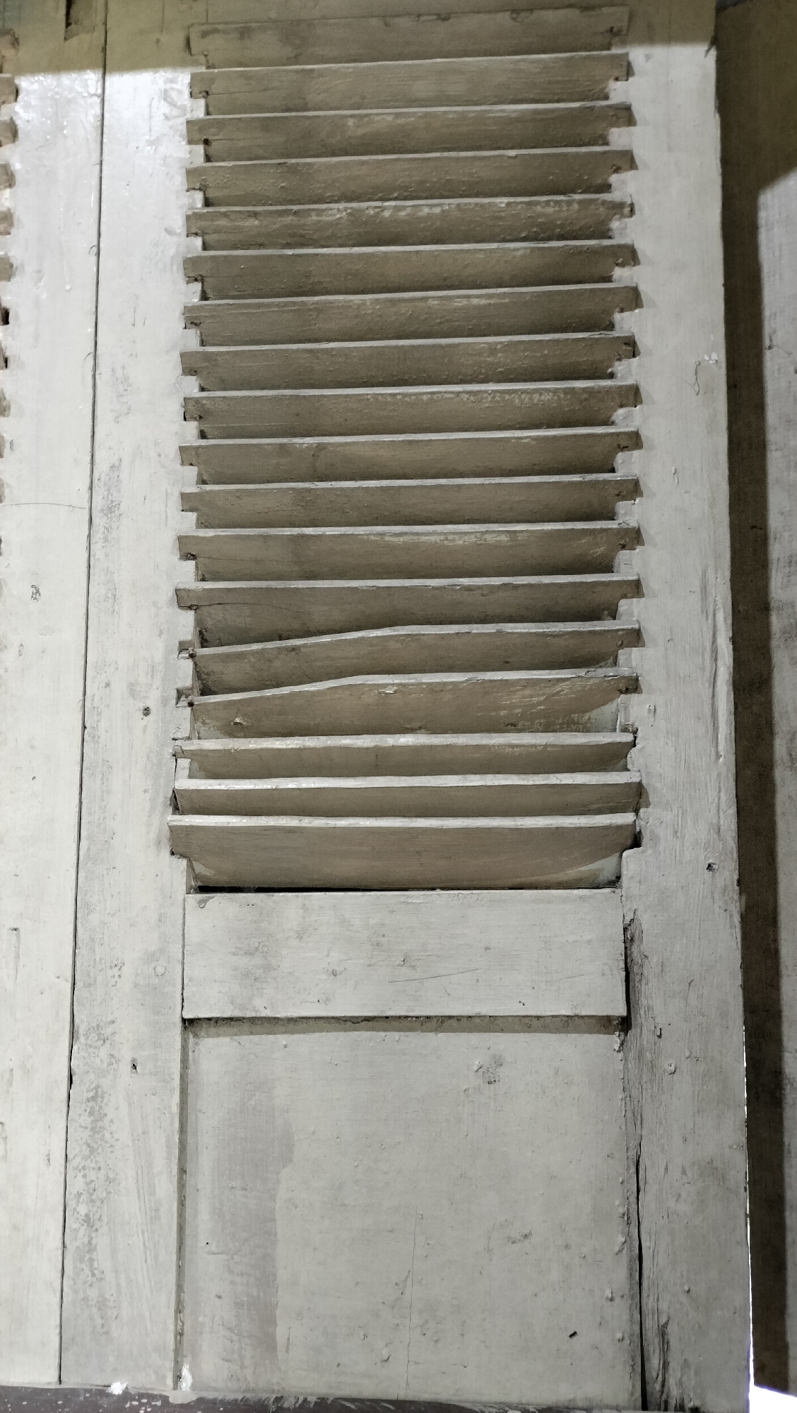This image shows a close-up view of a weathered, pale gray wooden shutter with horizontal slats. The shutter is attached to a wall or door frame, also made of aged wood. The slats are slightly uneven, with some appearing warped or broken, suggesting significant age and exposure to the elements. The texture of the wood is visible, with visible grain and imperfections like chips and cracks in the paint. The lighting suggests an indoor setting, possibly with light coming from the side, casting a shadow on the right side of the image. The overall impression is one of rustic simplicity and a sense of history. There is no text visible in the image. The location context is Tansaril, Indonesia.