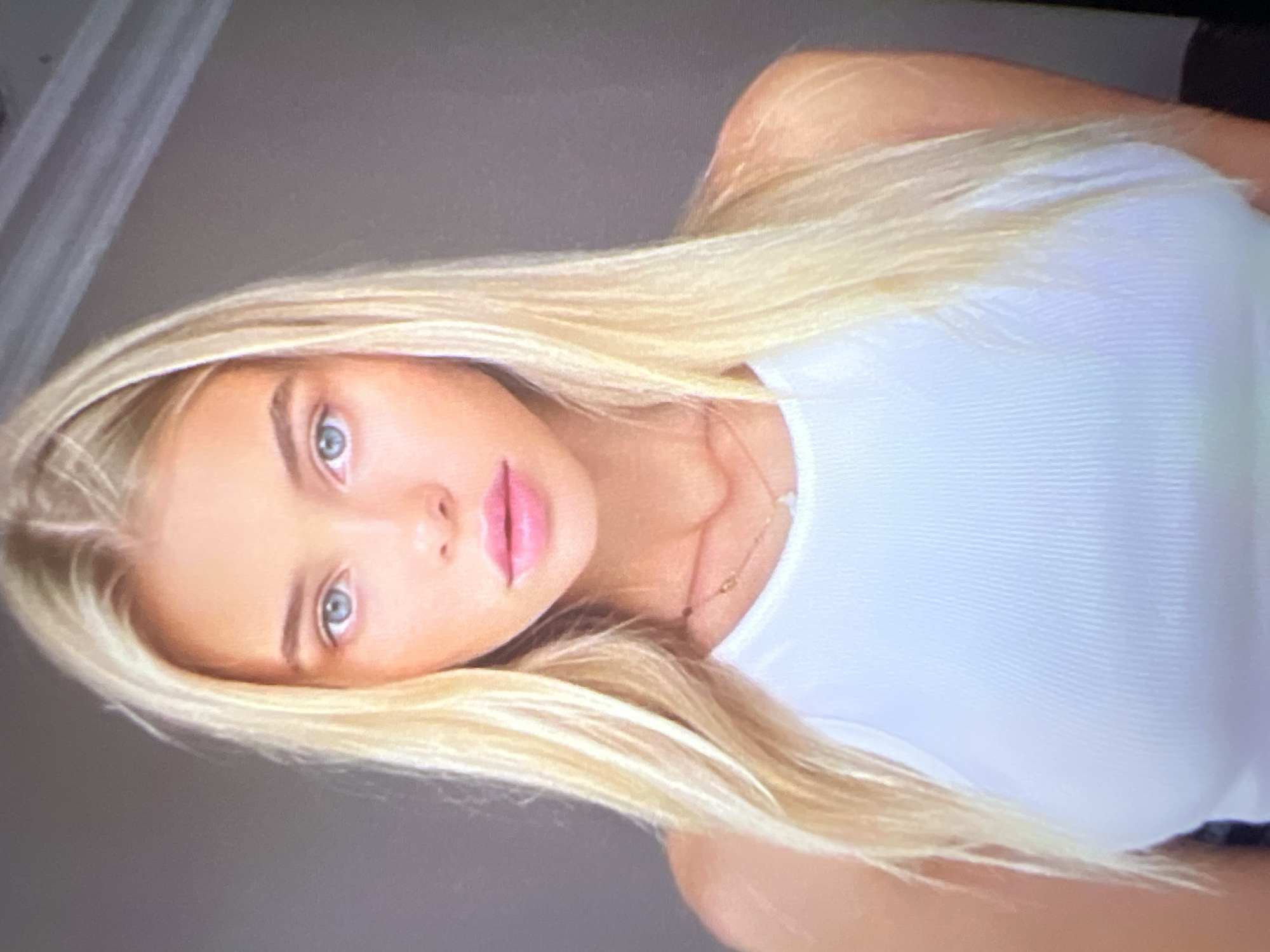 A young woman with blonde hair and blue eyes, wearing a white tank top and a delicate necklace, is photographed from the chest up. Her gaze is directed forward, and her lips are slightly parted. The background consists of a plain gray wall, with a sliver of a ceiling or upper wall visible in the top right corner. The lighting is soft and diffused, creating a gentle illumination on her face and upper body. This image does not contain any elements or information that would indicate a specific location within the city of Pyatigorsk, Russia.