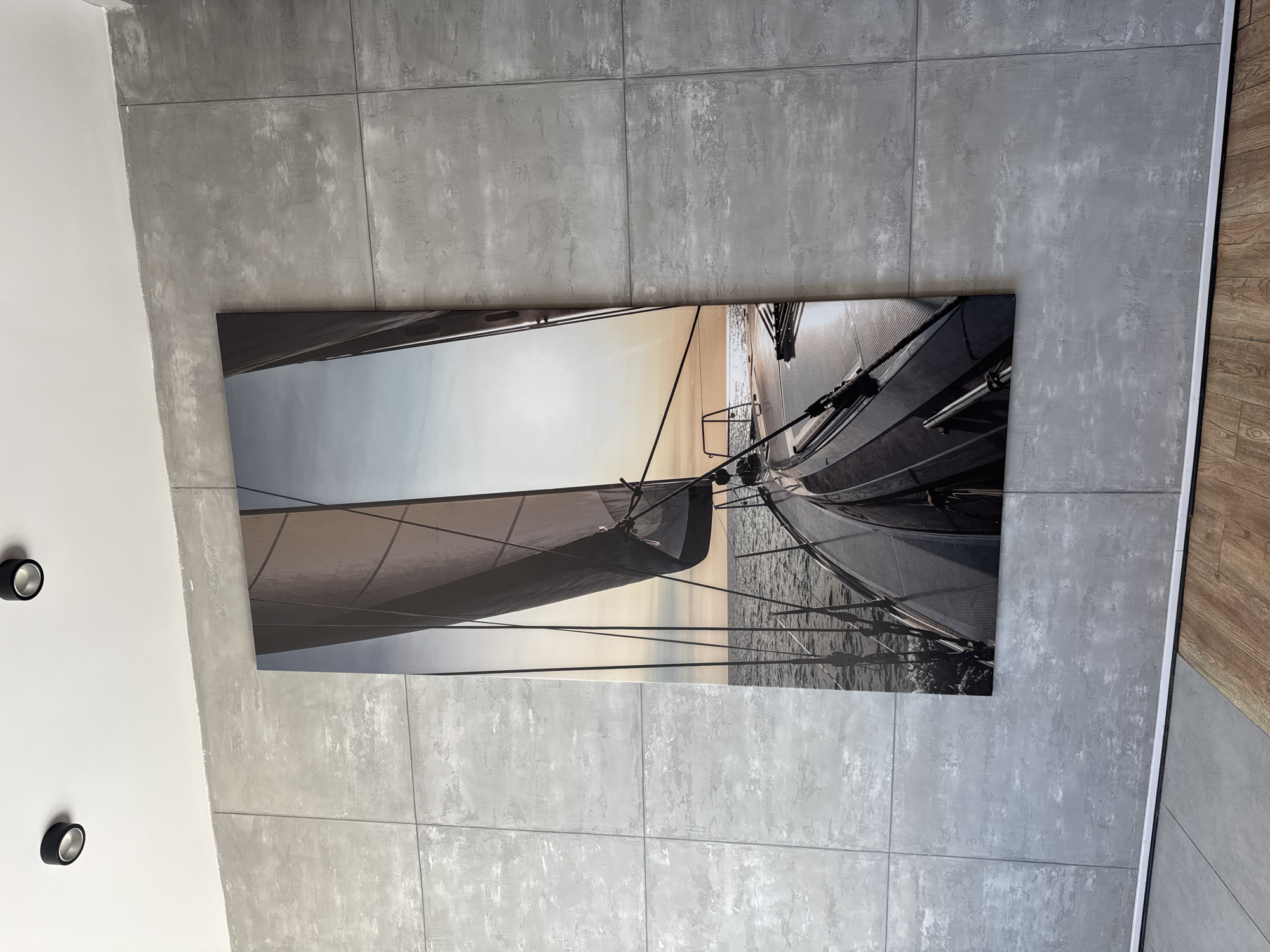 A large, vertically oriented framed canvas print depicting a sailboat at sea hangs on a grey tiled wall. The print shows a view from the deck of the sailboat looking forward, with a large sail in the foreground and the ocean stretching out to the horizon under a pale sky with the sun reflecting on the water. The wall is made of large, square concrete-look tiles. Two black cylindrical ceiling lights are visible in the upper portion of the image, mounted on a white ceiling. The lower part of the image shows a section of wooden flooring. The scene appears to be an interior space, possibly a living room or hallway, and there is no indication of the city of Saint Petersburg, Russia.