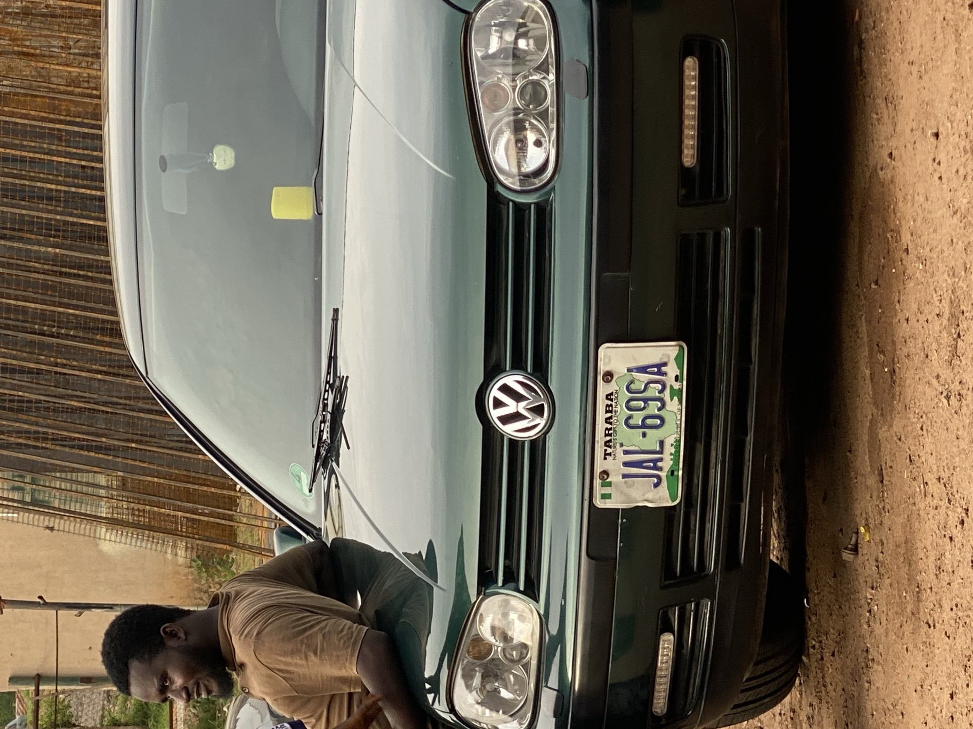 A man is leaning on the hood of a dark green Volkswagen Golf. The man is wearing a brown t-shirt and has a beard and short black hair. He is looking to his left, with his head tilted slightly. The car is parked on a dirt road, with a fence made of metal bars in the background. The license plate of the car is visible and reads "JAL-69SA TARABA NATURE'S GIFT TO THE NATION". The location is implied to be Jalingo, Nigeria, given the license plate. A yellow object and a small pendant shaped like an apple are hanging from the windshield. There are also LED lights on the bumper of the car. The ground is covered in loose dirt and small rocks.