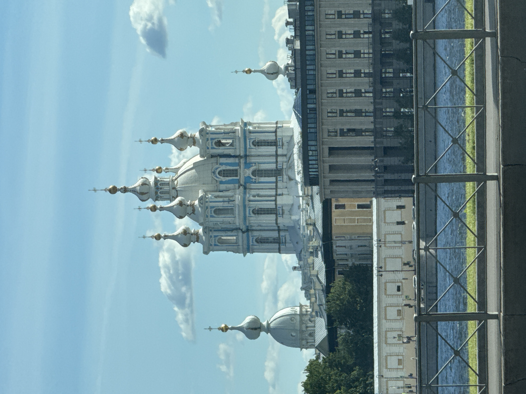 This is an outdoor, eye-level shot of the Smolny Cathedral in Saint Petersburg, Russia. The image is oriented vertically, with the sky and the upper portions of the cathedral appearing at the bottom of the frame, and the ground and water at the top.
The primary subject is the Smolny Cathedral, a striking blue and white Baroque building with multiple onion domes topped with gold crosses. One large dome is prominent, and several smaller ones are visible. To the right of the cathedral, a modern, multi-story building with numerous windows stands. Between these two structures, a body of water, likely a canal or river, is visible, bordered by a patch of green grass and a metal fence with a diamond pattern. Trees can be seen in the background. The sky is a bright, clear blue with a few white, puffy clouds. The lighting suggests a sunny day, likely mid-day given the brightness.
No people are visible in the image. The scene depicts an architectural landmark within an urban setting. The overall impression is one of grandeur and historical significance juxtaposed with modern architecture and a natural element of water.