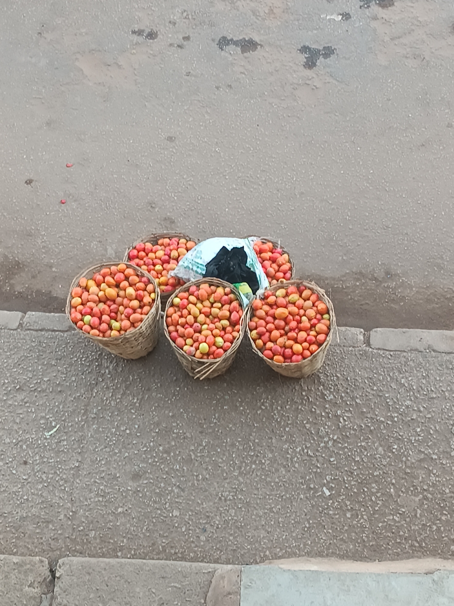 The image shows five baskets and a plastic bag filled with red and yellow tomatoes on the edge of a paved road. The location is Bukuru, Nigeria.