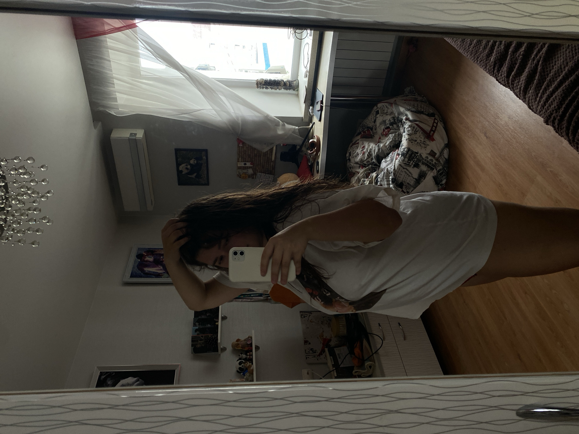 The image is a mirror selfie taken indoors, likely in a bedroom or living area. A young woman with dark hair is posing in front of the mirror, holding her phone to take the photo. She is wearing a loose-fitting white t-shirt. The room appears to be furnished with a desk near a window, a beanbag chair, and shelves with various decorative items and books. The lighting suggests it is daytime. The window shows a glimpse of the outside, with some buildings visible, though the exact location is not identifiable. There is no information within the image to suggest the city of Perm, Russia.