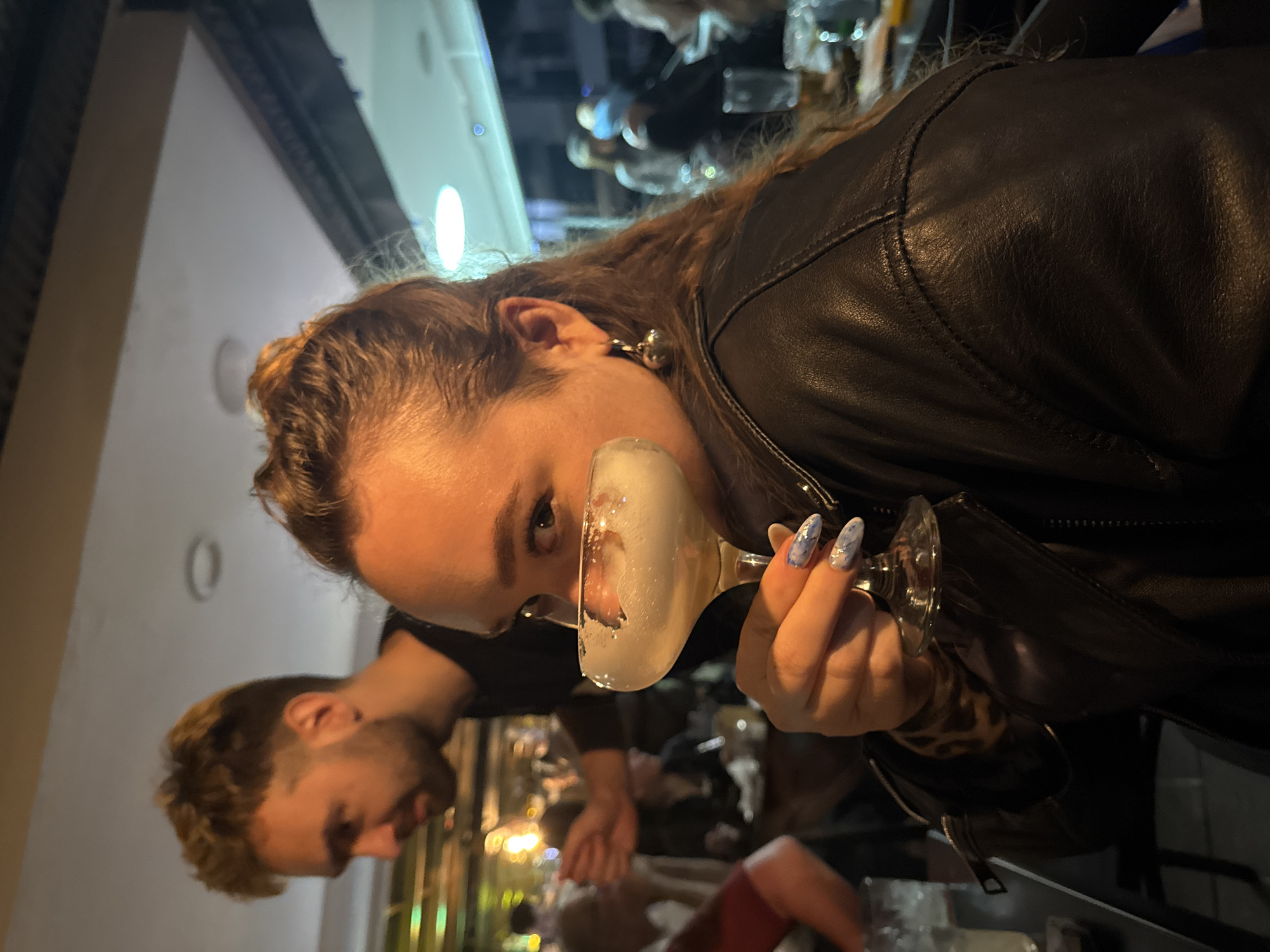 An eye-level photograph, taken at a Dutch angle, captures a woman holding a cocktail glass in what appears to be a dimly lit bar or restaurant in Ramat Gan, Israel. The woman, with long, wavy brown hair styled back, is the primary subject, positioned in the foreground and looking directly over the rim of her glass. Her expression is calm and observant. She wears a black leather jacket over a garment with a leopard print pattern, visible on her right arm. Her fingernails are notably long and adorned with a distinctive blue and white marbled or patterned design. She holds a stemmed glass filled with a light-colored, frothy drink that glistens under the ambient lighting. Small, silver-toned earrings are visible on her left ear.
In the blurred mid-ground, slightly below and behind the woman, a man is partially visible, primarily his profile. He has short, curly light brown hair and a beard or stubble. He appears to be looking towards the left side of the frame, away from the camera. The background is dark and out of focus, showing indistinct lights and shapes consistent with an indoor evening setting. The overall mood is casual and social. No visible text is present in the image.