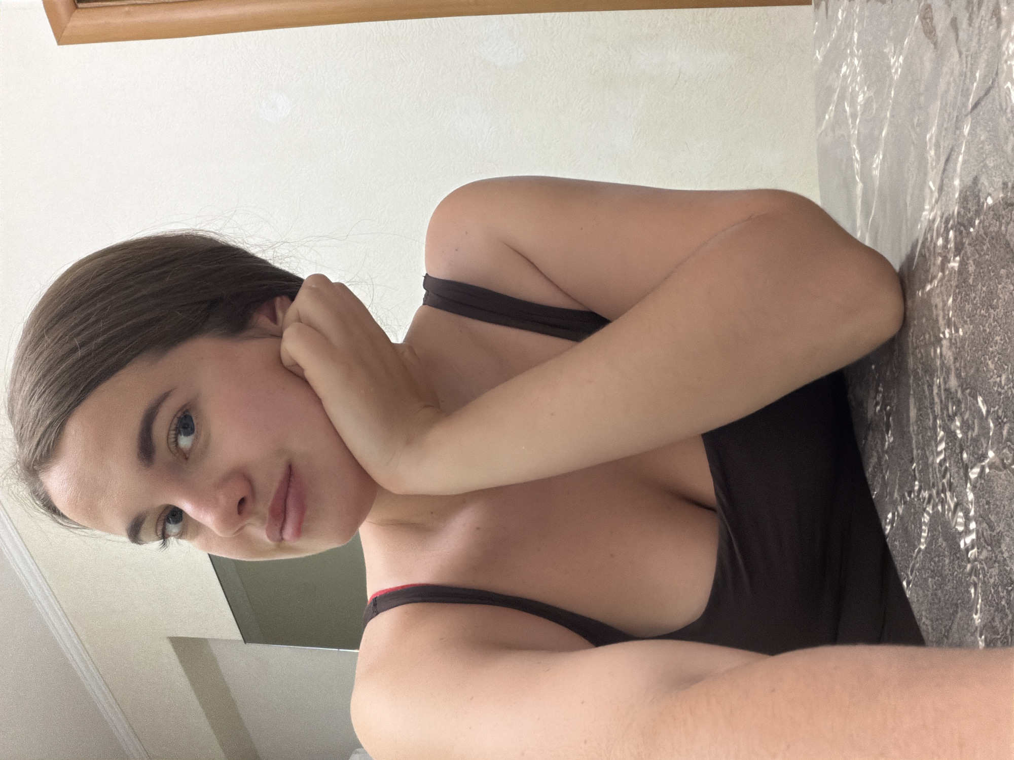 A young woman poses for a close-up shot in an indoor setting. She has long, dark hair and fair skin. She is wearing a dark-colored tank top with a red trim along the straps. Her face is turned towards the camera, and she is resting her cheek on her hand. The background includes a wall, a wooden trim, and a patterned surface that could be a countertop or table. The location is unknown.