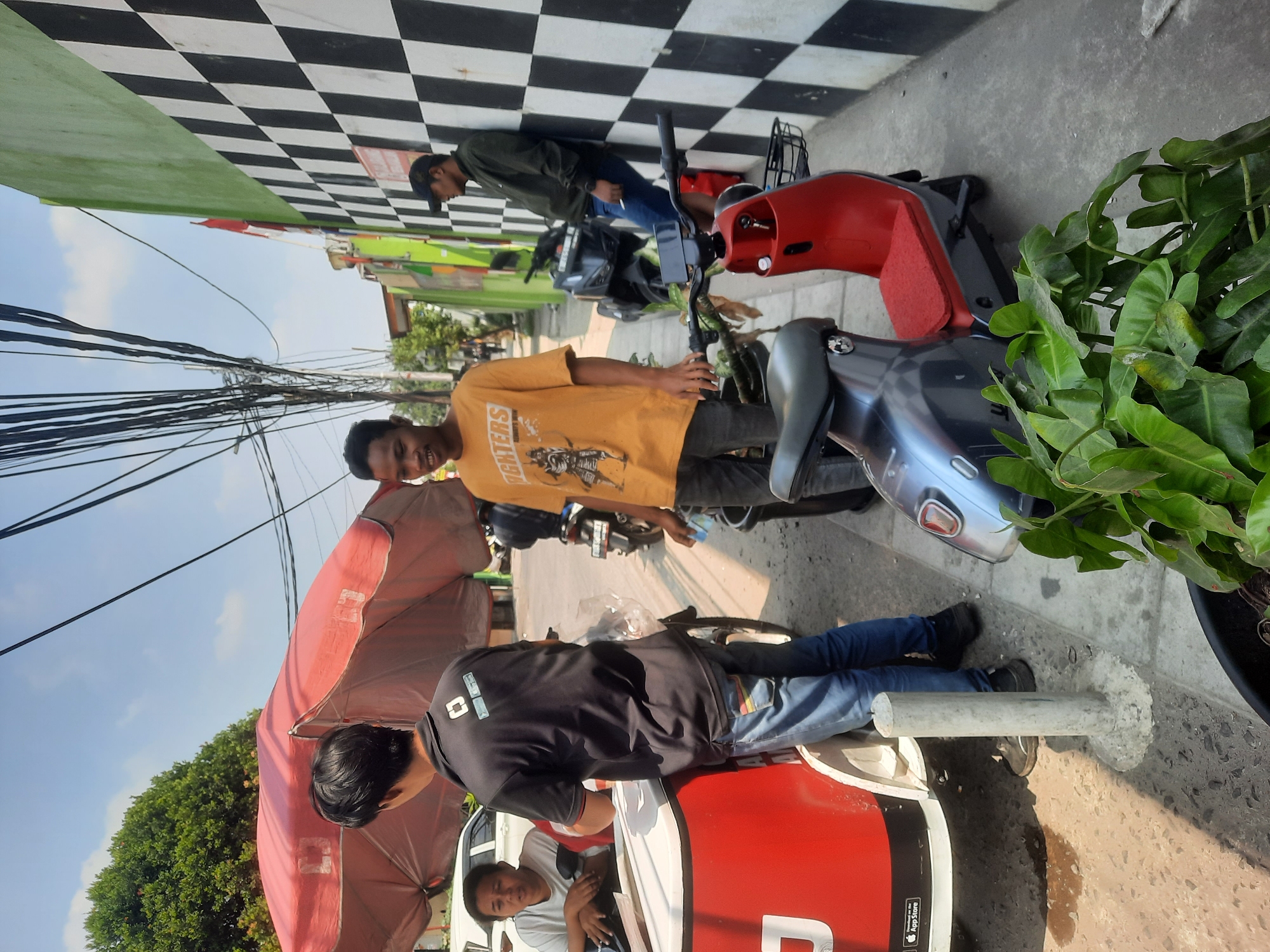 The image depicts a street scene in North Jakarta, Indonesia, on a bright, sunny day. In the foreground, a person wearing an orange t-shirt stands near a red and gray electric scooter. To their left, another person in a black t-shirt is interacting with a white and red cart, possibly a food or beverage vendor. A person with short hair is visible inside the cart. In the background, a man in a dark jacket leans against a wall adorned with a black and white checkered pattern. Several motorcycles are parked along the street, and overhead power lines crisscross the sky. Lush green foliage is present in the lower right corner, suggesting a touch of nature amidst the urban environment. The overall atmosphere appears to be casual and bustling.