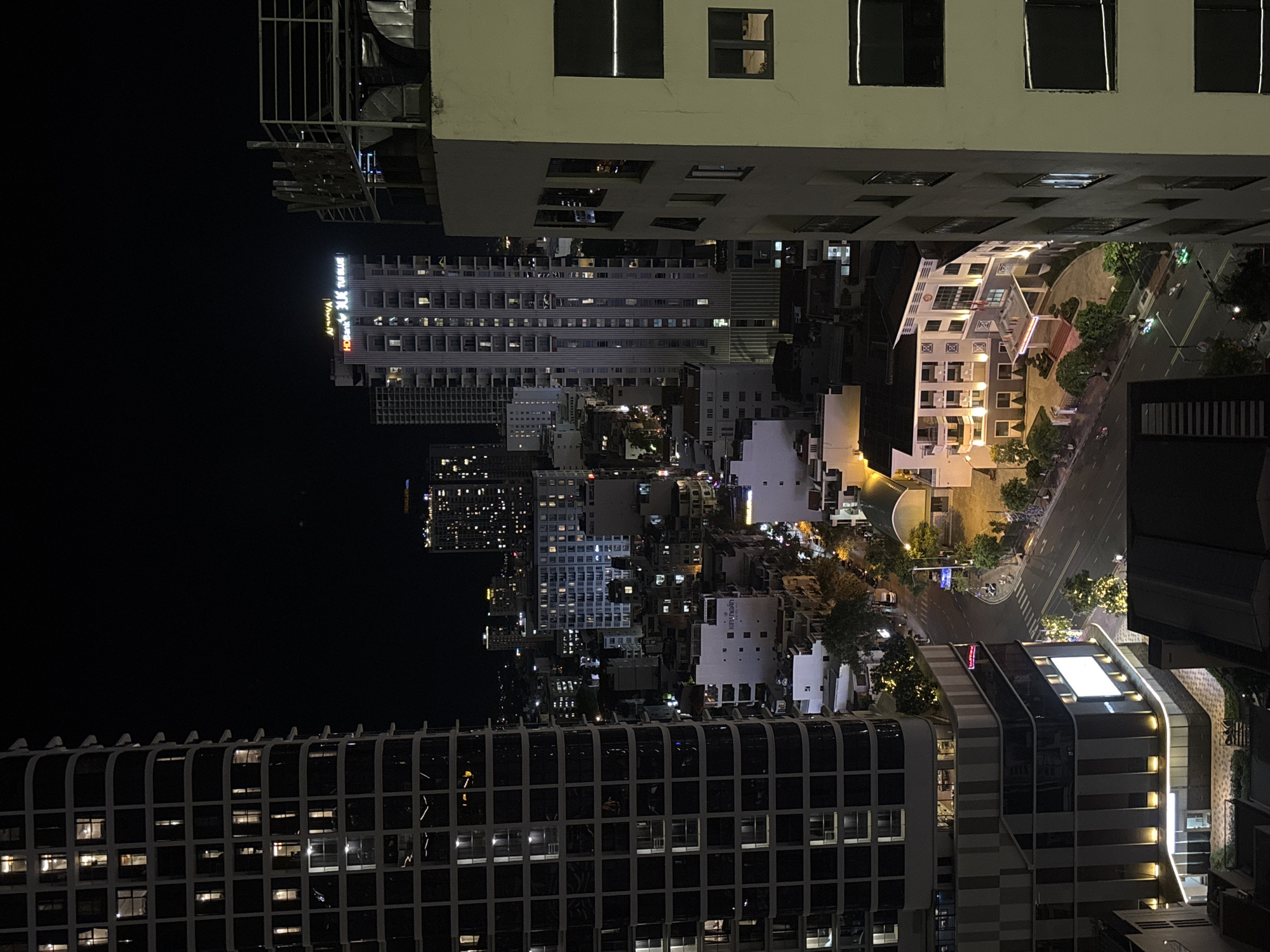 This is a nighttime aerial view of a city street in Nha Trang Ward, Vietnam, dominated by towering buildings and streetlights. The perspective is from a high vantage point, looking down on an intersection with traffic and pedestrians.
On the left, a large, modern skyscraper with a dark facade and numerous illuminated windows dominates the frame. Its curved design and stacked geometric sections are visible. Across the street, a lighter-colored, multi-story building with a prominent illuminated sign at its base is visible. This building has a modern architectural style with balconies and glass facades.
Further into the background, more city buildings are visible, some illuminated and others dark, creating a dense urban landscape. The street itself is paved and appears to be a busy thoroughfare with vehicles, including cars and motorcycles, moving along it. Streetlights and lights from the buildings cast a warm glow on the scene. Trees line the sides of the street in some areas, adding greenery to the urban environment.
A few people can be seen on the sidewalks, and some are riding motorcycles on the street. The overall impression is a bustling city at night, with a mix of modern and older architecture. The sky is dark, indicating nighttime. The image appears to be taken from a significant height, offering a broad view of the urban setting.