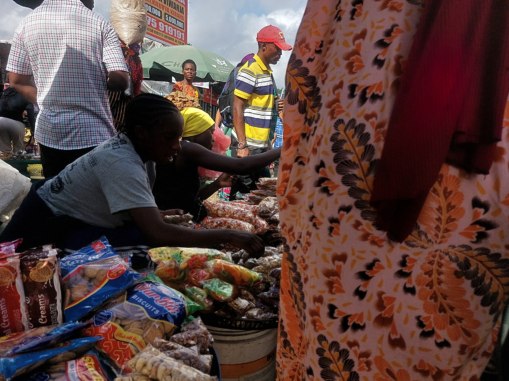 This bustling scene depicts an outdoor market in Dar es-Salaam, Tanzania, under a bright, cloudy sky. The foreground is dominated by a display of packaged snacks, including various biscuits and nuts, suggesting a vibrant trade in everyday food items. Brands such as "Star Biscuits" and "Var-Bit Biscuits" are visible on the colorful packaging.
Several individuals populate the market. On the left, a person in a grey t-shirt with "Kaphics" and "PRINTING" visible text, possibly a vendor, leans over a stall, arranging goods. Another young person with a yellow headscarf is also attentive to the products. In the mid-ground, a man wearing a yellow and blue striped shirt and a red baseball cap stands, while behind him, another man in a plaid shirt is seen from the back. To the left, a woman in an orange patterned top sits behind a stall. On the far right, part of a person wearing a distinct floral-patterned dress in hues of orange, black, and white is visible, with a maroon fabric draped nearby.
A large white sack is balanced on someone's head in the background, a common sight in busy markets. Above the stalls, a large sign with text "KILUVYA, KIBAHA", "IWANJA", numbers like "5,000,000" and "919191" (likely a phone number) and "JOGO KIDOGO UKUBWA NI 2020" provides local context. A green market umbrella offers shade in the background. The overall atmosphere is lively, indicative of daily commerce and community interaction.