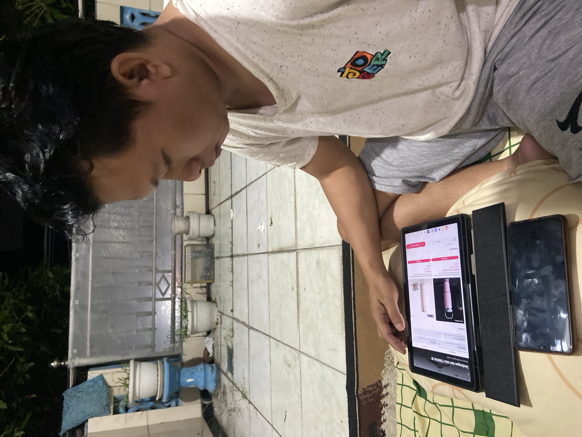 The image shows a person sitting on a floor with a patterned blanket. They are looking down at a tablet and a smartphone, both of which are placed on the blanket. The person is wearing a white t-shirt with a colorful graphic on the chest and gray shorts. The tablet displays a webpage with product images and text, likely from an online shopping site. Next to the tablet is a dark-colored smartphone. The scene appears to be indoors, possibly in a living area, with tiled flooring visible in the background. The lighting suggests it could be evening or nighttime. The person's posture and focus on the devices indicate they are engaged in using them, perhaps for browsing or shopping. The overall atmosphere is casual and focused. The location context is Gelam, Indonesia.