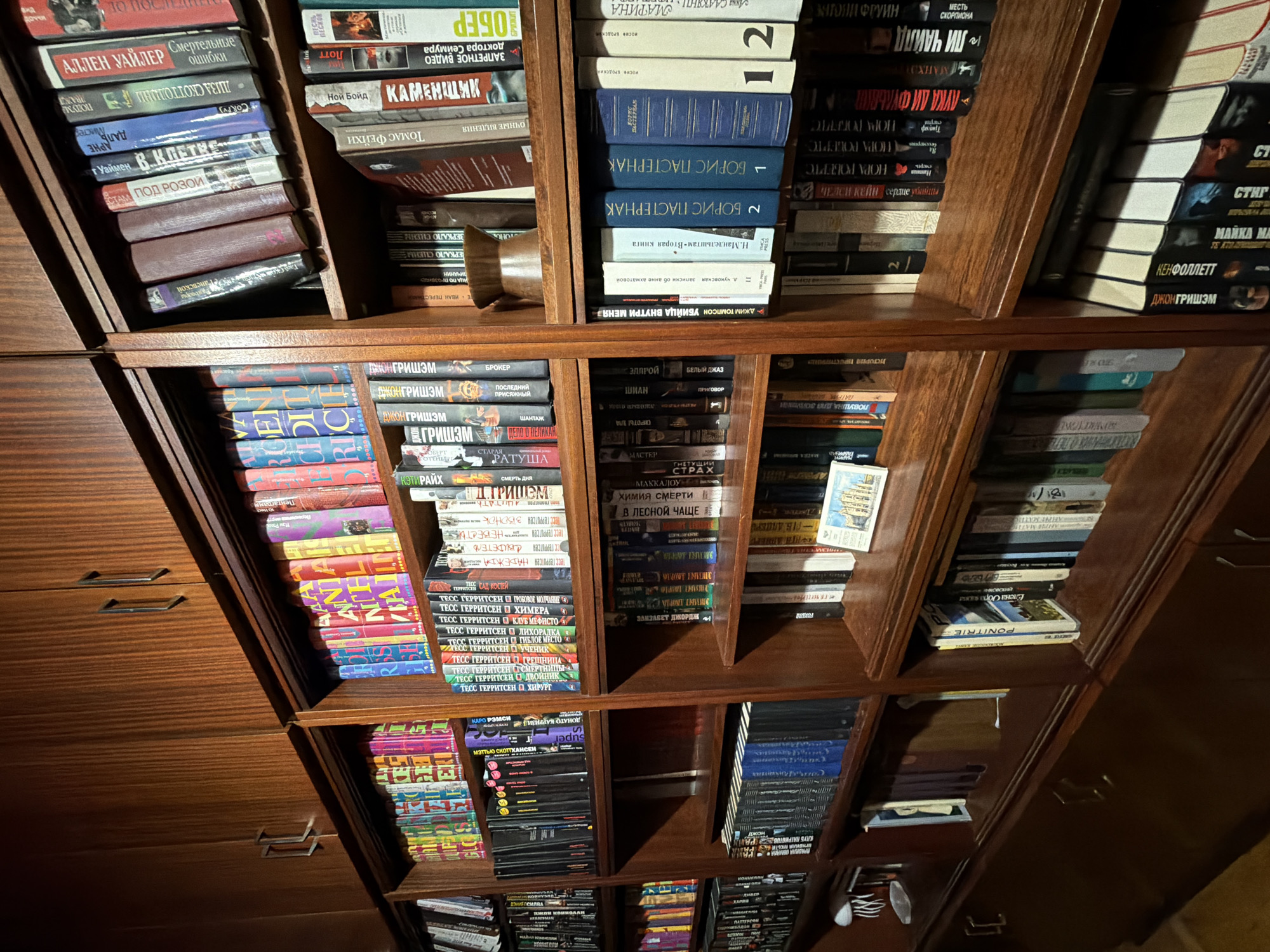 The image shows a dark brown wooden bookshelf filled with various books. The books are arranged in stacks and rows across different shelves. The titles on the spines of some books are visible, including authors like John Grisham, Tess Gerritsen, and Boris Pasternak. The books are primarily in Russian, with some English titles interspersed. The bookshelf itself appears to be part of a larger cabinet, with drawers visible on the left side. The lighting is somewhat dim, highlighting the textures of the wood and the books.