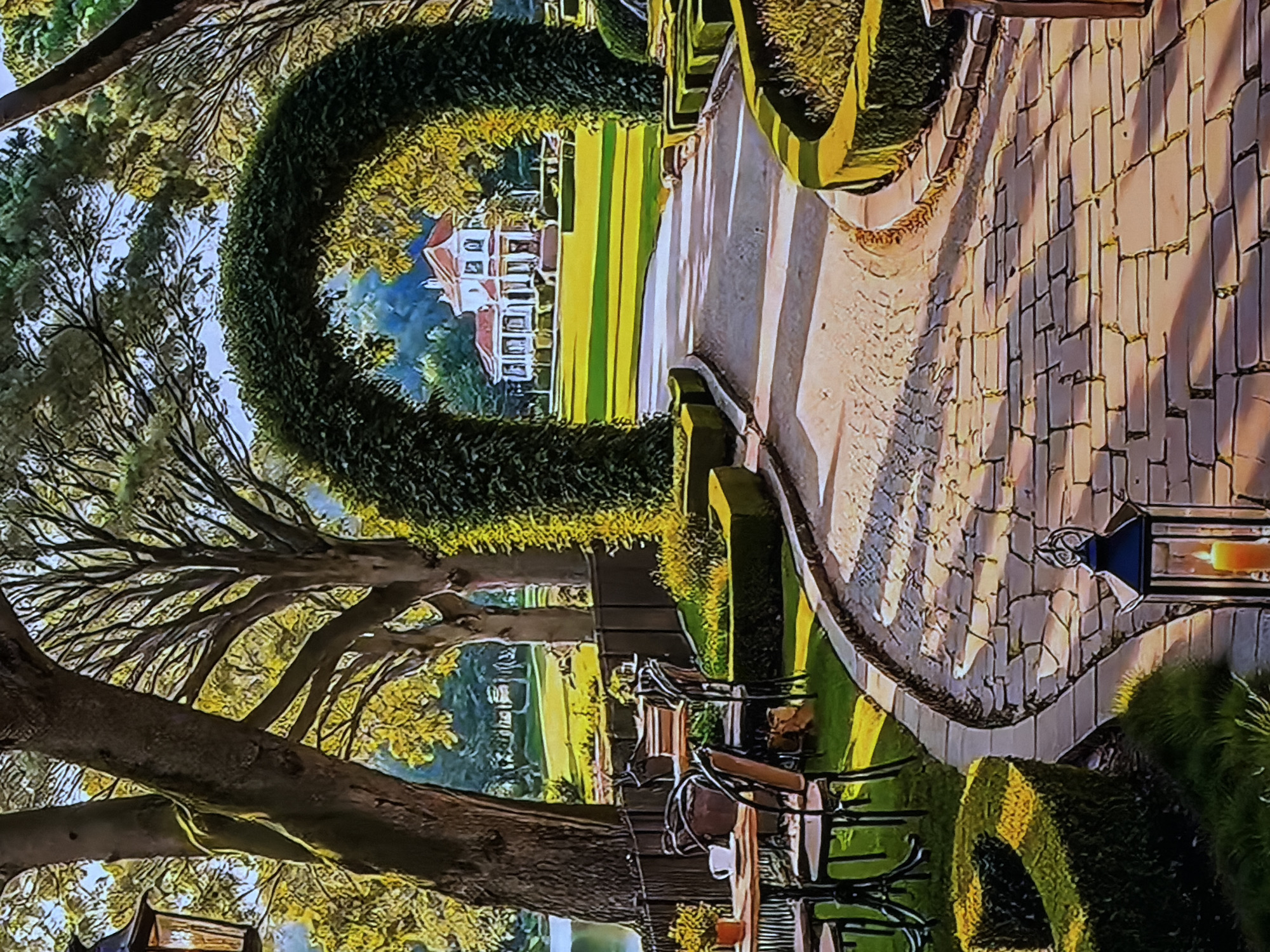 The image displays a picturesque and well-maintained garden scene, likely part of an estate, resort, or large park. The perspective is slightly elevated, looking down a curving pathway towards a distant building.
In the foreground and mid-ground, a wide, paved pathway constructed from light-colored rectangular blocks meanders through the landscape. The pathway is bathed in warm, directional sunlight, which casts long shadows across its surface and the surrounding areas, suggesting either early morning or late afternoon.
A prominent feature is a large, meticulously trimmed U-shaped green hedge or topiary that forms an archway in the mid-ground. Beyond this hedge, an expansive, striped green lawn stretches towards a grand, multi-story white building with distinctive red roofs, positioned centrally in the background. The building appears to be a mansion or a stately home.
To the left of the scene, large, mature trees with visible branches and foliage in shades of green and yellow frame the view. On the right side, closer to the viewer, an outdoor patio area is suggested by glimpses of tables and chairs, alongside manicured hedges. A decorative dark metal lantern, containing a visible light source, possibly a candle, stands near the path's edge on the right.
The overall atmosphere is serene and elegant, characterized by lush greenery, carefully maintained landscaping, and the warm glow of natural light. No specific human activity is visible, but the setting suggests a place for relaxation or formal gatherings. There is no visible text in the image. The provided location context for this scene is Sofiivska Borshchahivka, Ukraine.
